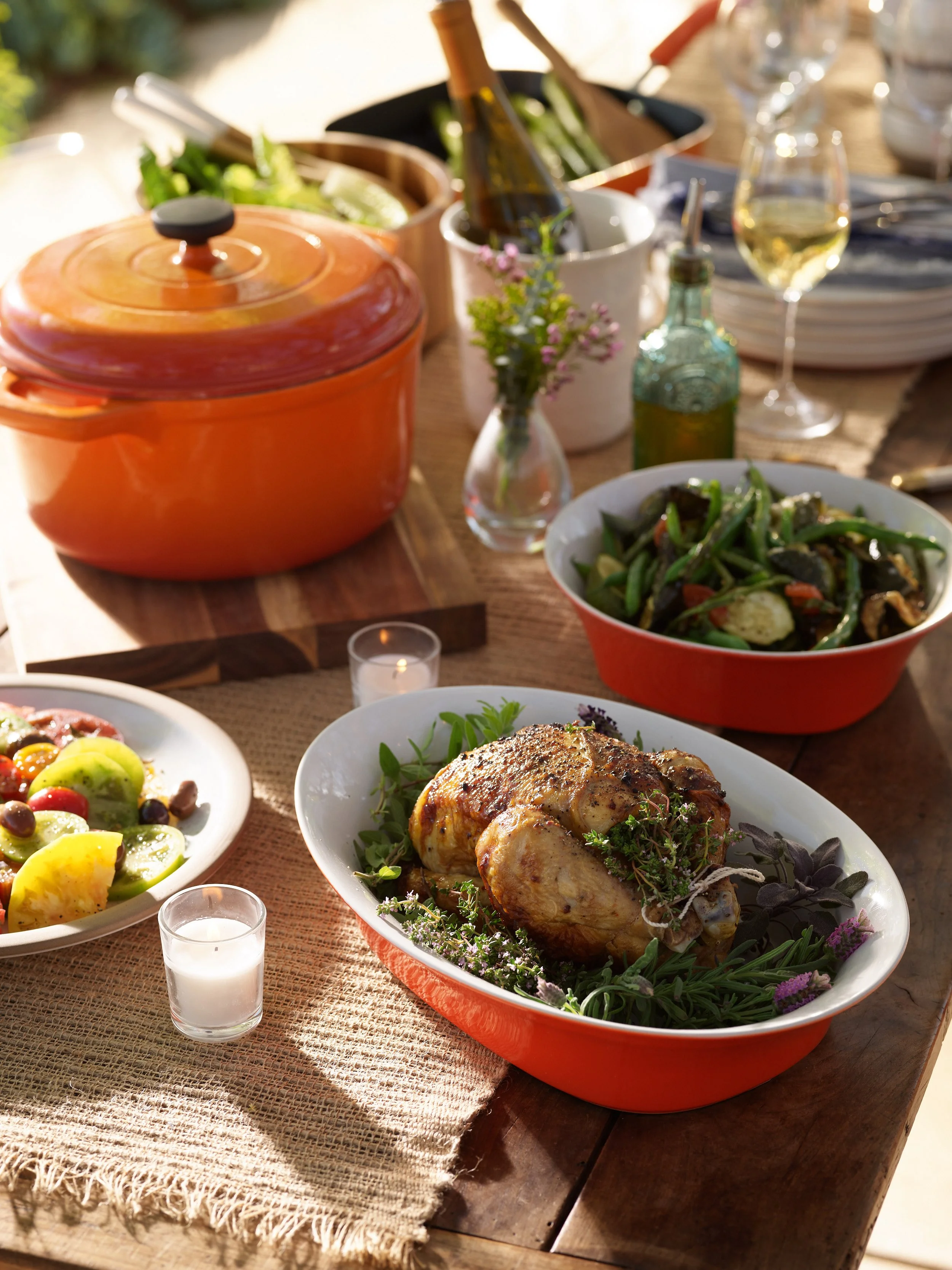 A table set with a roasted chicken dish garnished with herbs, a vegetable salad, a bowl of green beans and vegetables, a plate of tomato and cucumber salad, wine glasses, a small bottle of olive oil, a small vase with flowers, and candles, indicating