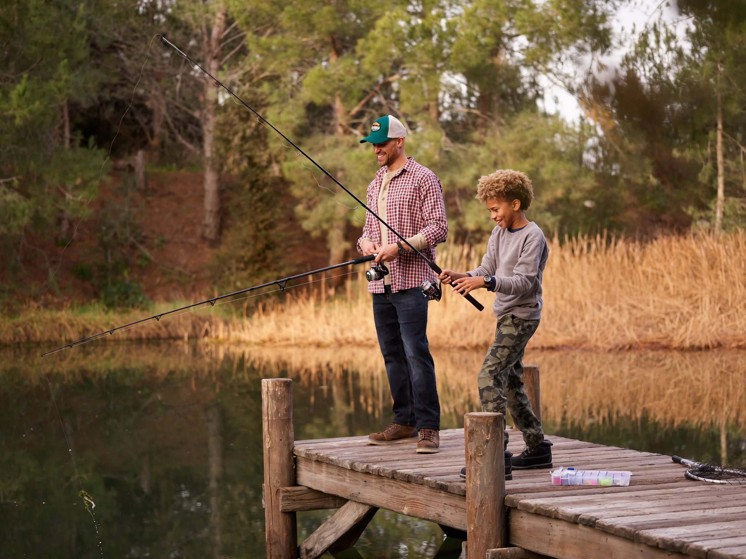 A man and boy fishing on a wooden dock by a lake surrounded by trees and tall grass.