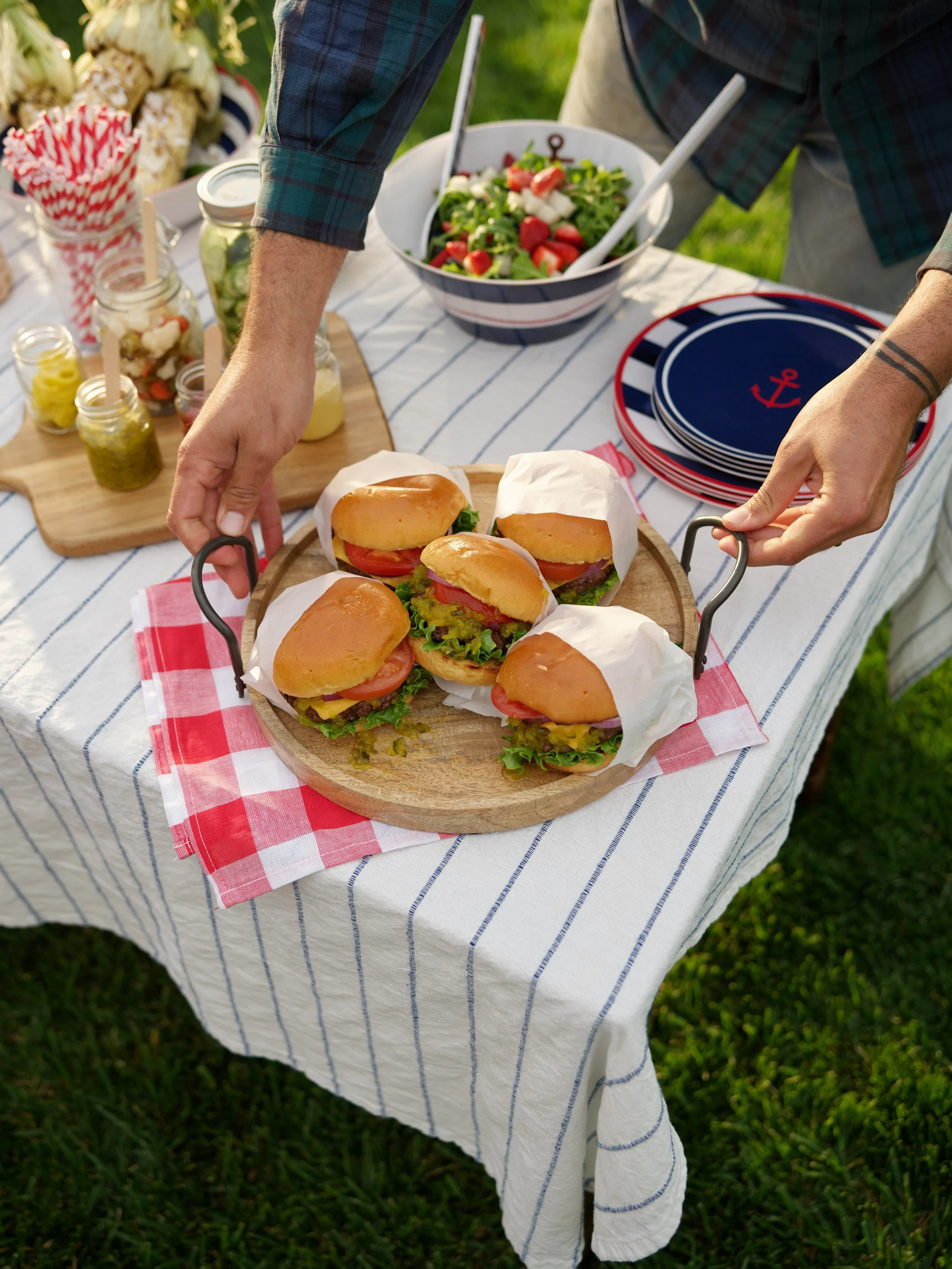 A person placing four small sandwiches with lettuce, tomato, and meat on a wooden tray at a picnic table with a red and white checkered napkin. The table has a blue and white striped tablecloth and other food including a salad and jars of pickles and