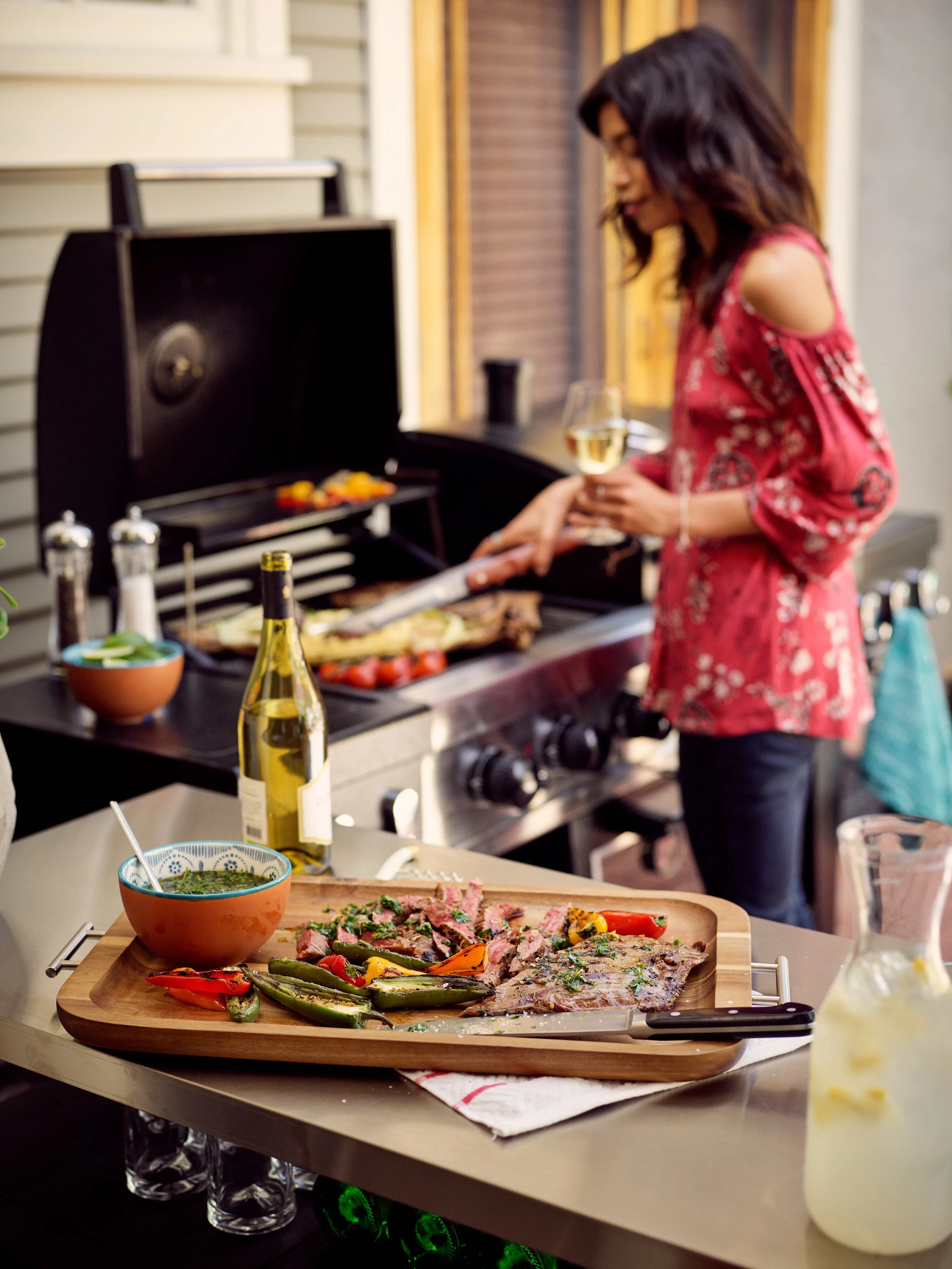 A woman wearing a pink floral top is grilling food on a barbecue grill while holding a glass of white wine. On a table in the foreground, there is a wooden serving tray with grilled vegetables, sliced cooked meat, a bowl of green sauce, and an open b