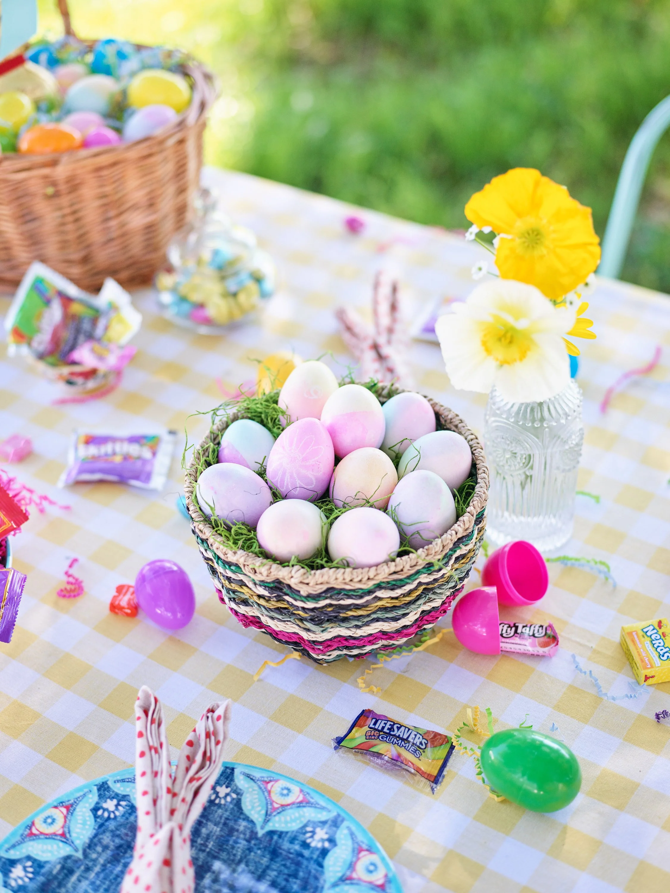 Easter table with a basket of pastel-colored decorated eggs, candy, and a vase of yellow and white flowers on a yellow checkered tablecloth outdoors.