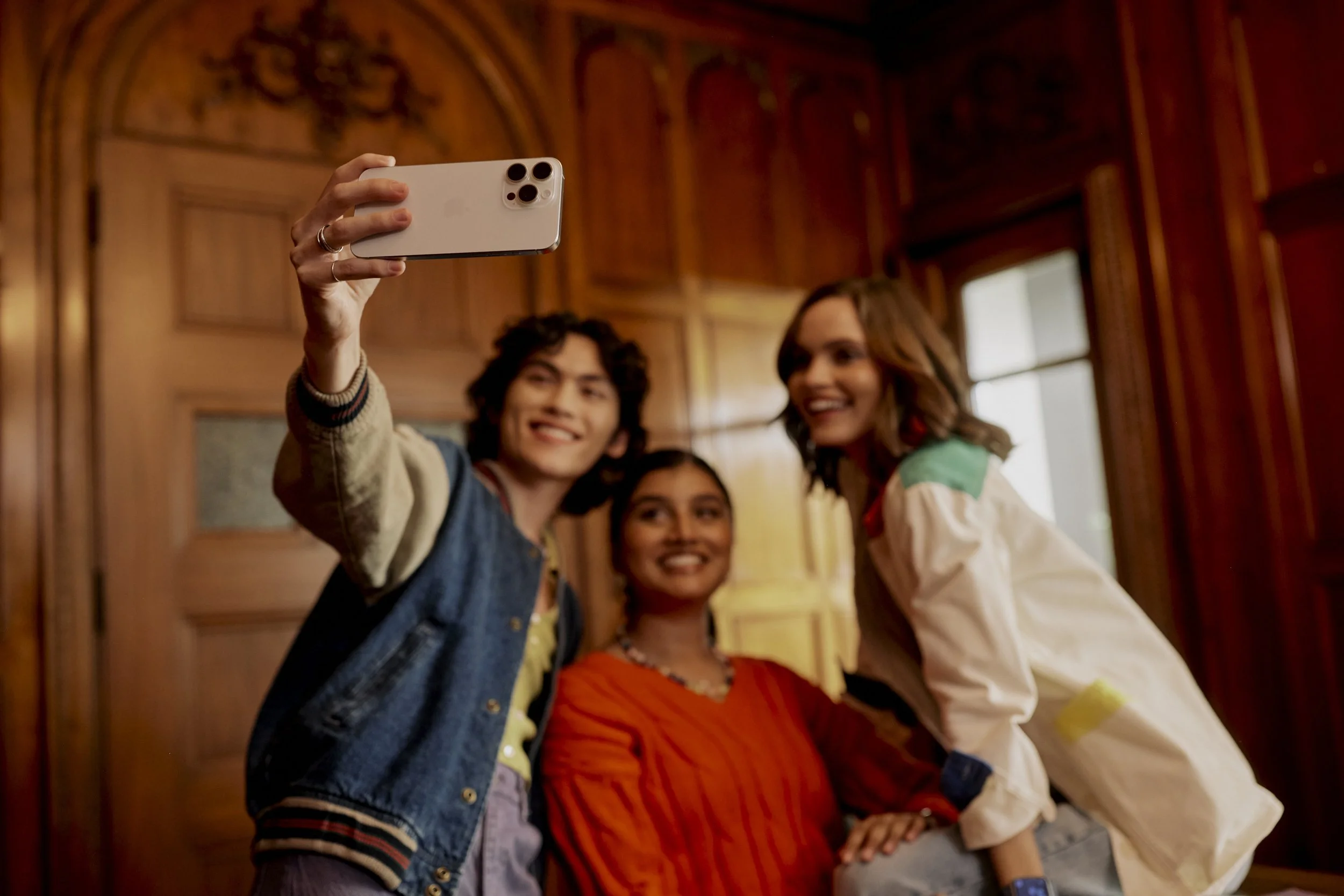 Three young women taking a selfie indoors, smiling, with wooden walls and a window in the background.