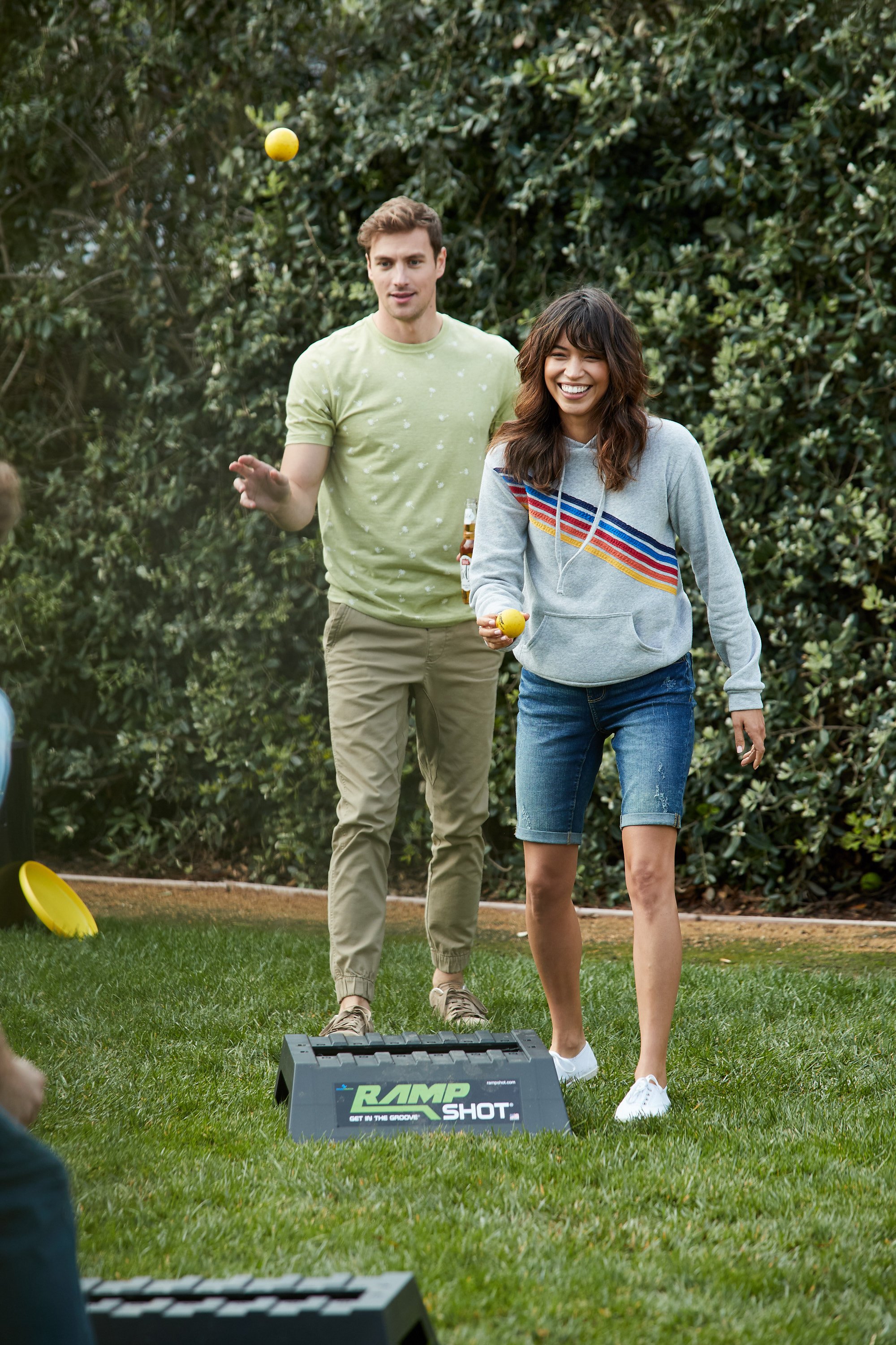 Two young adults playing cornhole outdoors, with one woman in a gray sweatshirt and denim shorts tossing a yellow bean bag, and a man in a light green t-shirt watching, with trees in the background.