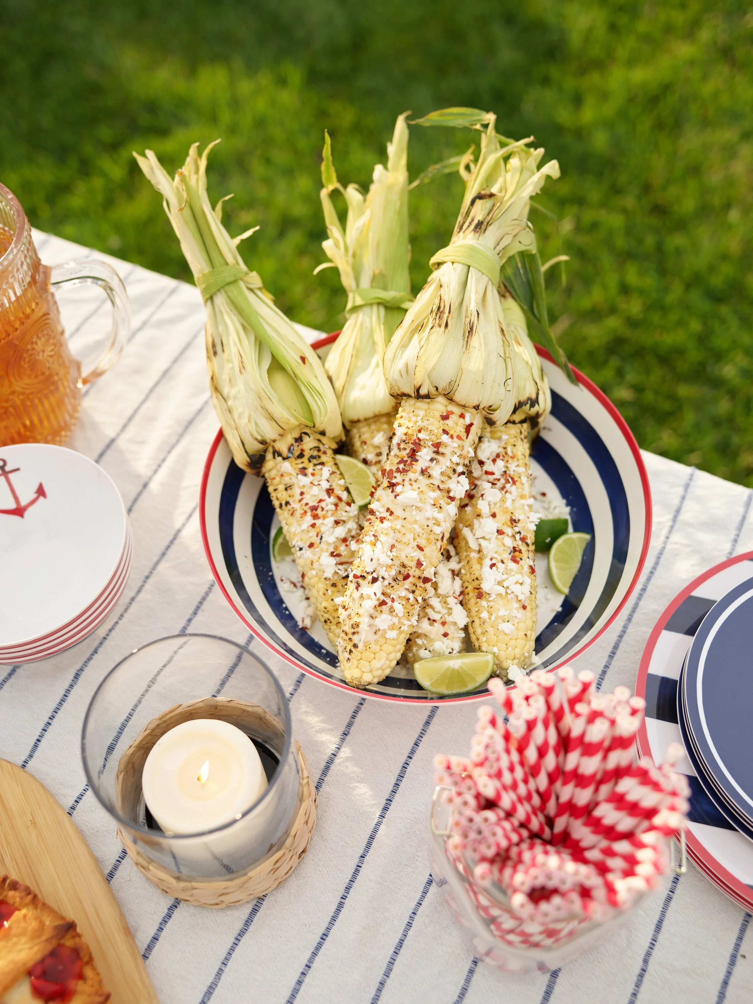 Grilled corn on the cob with seasoning and lime wedges on a striped plate at a picnic table with a candle, red-striped straws, and other dishes visible.