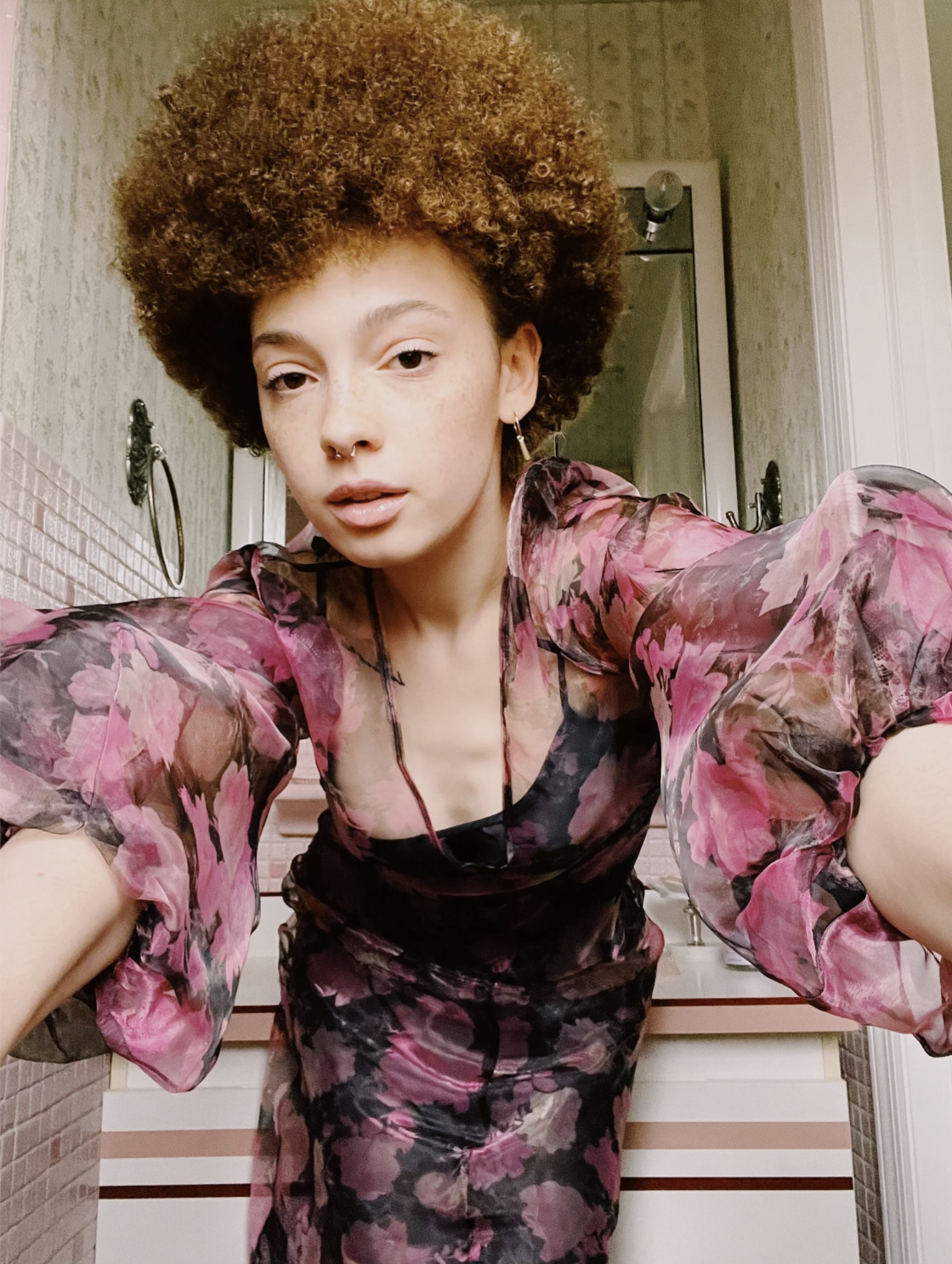 A person with short curly hair, wearing a floral blouse, leaning forward towards the camera in a bathroom with pink and white striped cabinets in the background.