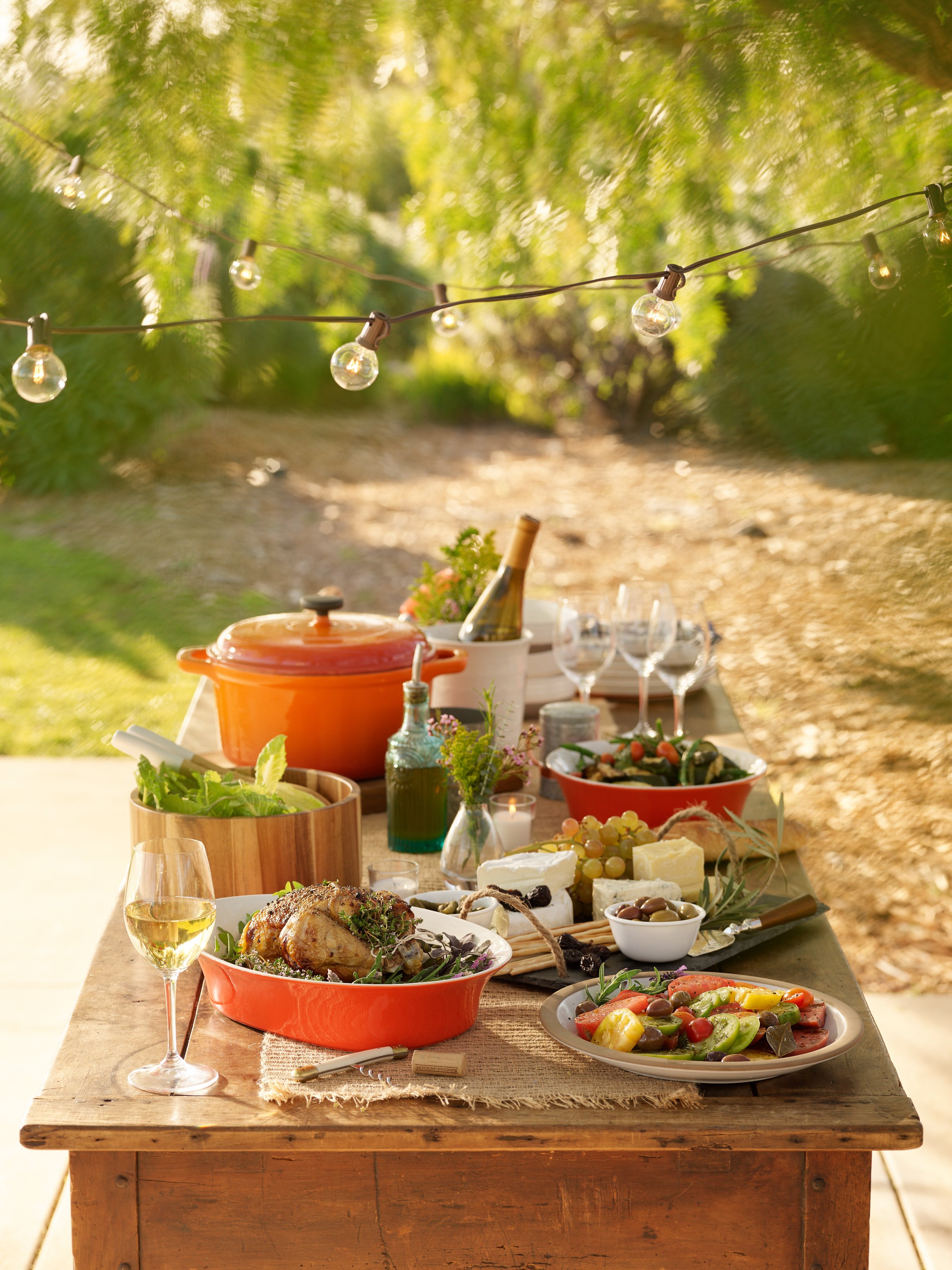 An outdoor table set with a variety of dishes including a roasted chicken, cheese platter, salad, wine, and decorative items, under string lights on a sunny day.