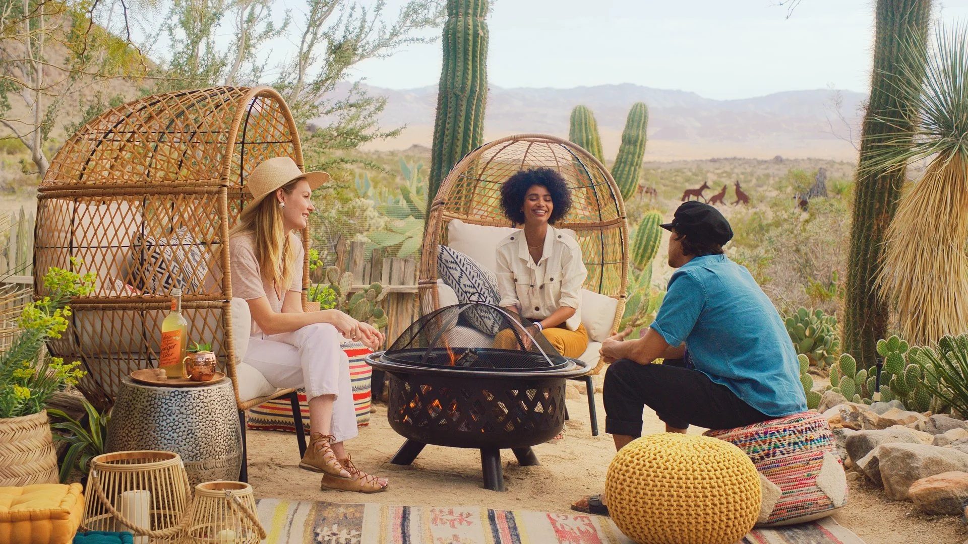 Three friends sitting around a fire pit with desert and cactus landscape in the background, two inside woven pod chairs and one on a decorative cushion on the ground, enjoying drinks and conversation.