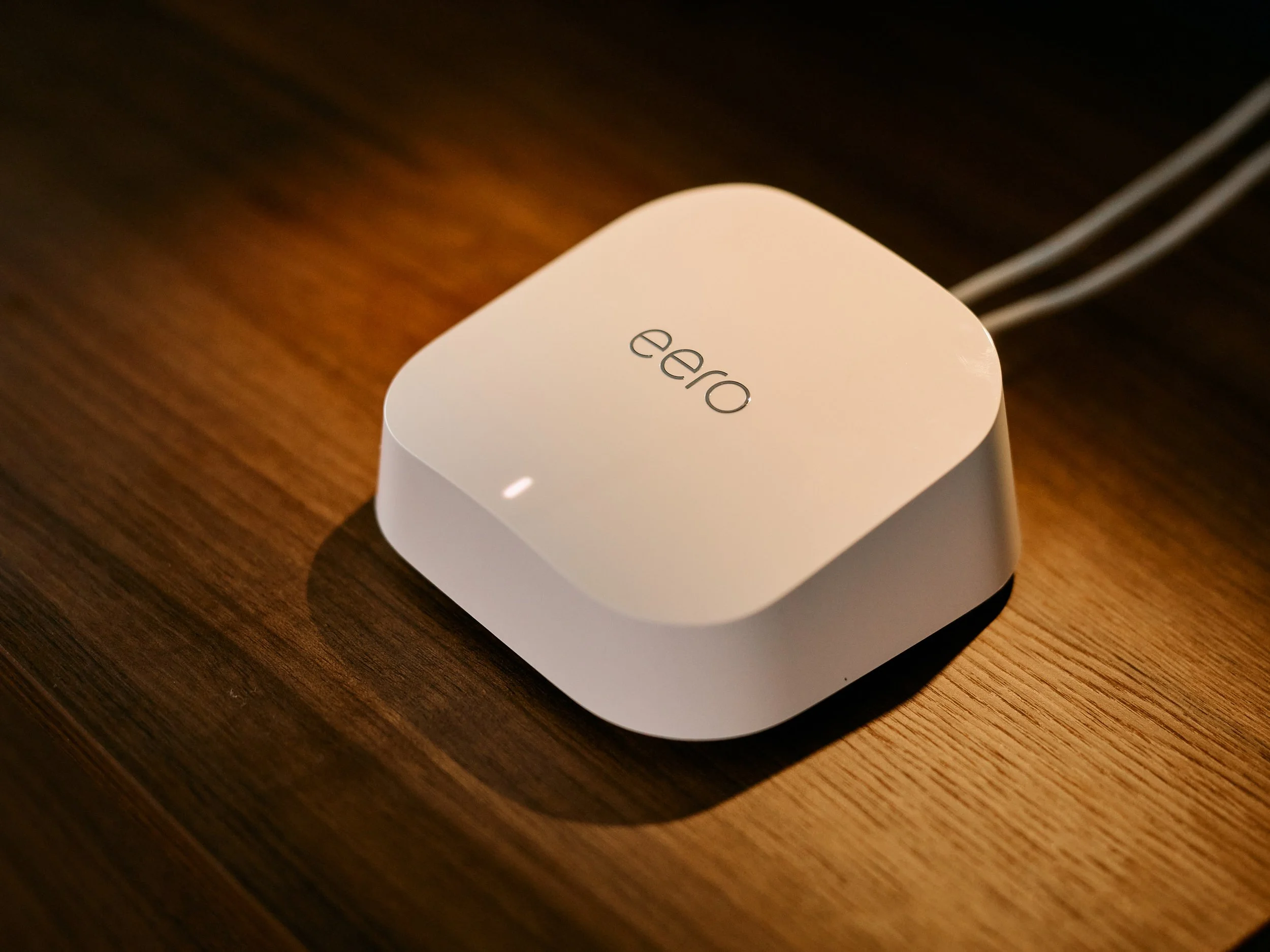 A white electronic device with the word 'eero' on top, placed on a wooden surface, connected by two white cables.
