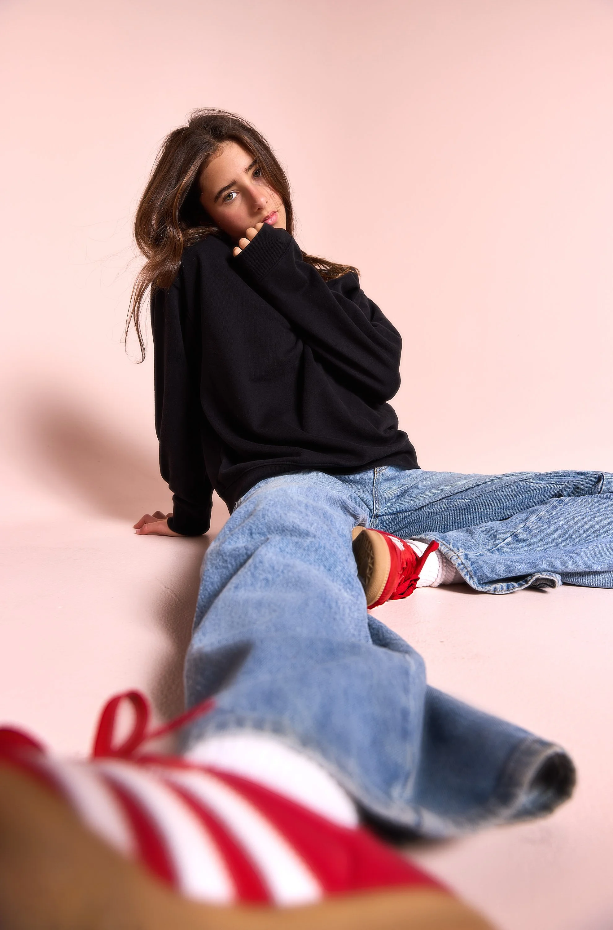 Young woman with long brown hair wearing a black hoodie, light blue jeans, and red sneakers, sitting on a light pink background with a relaxed pose and a thoughtful expression.