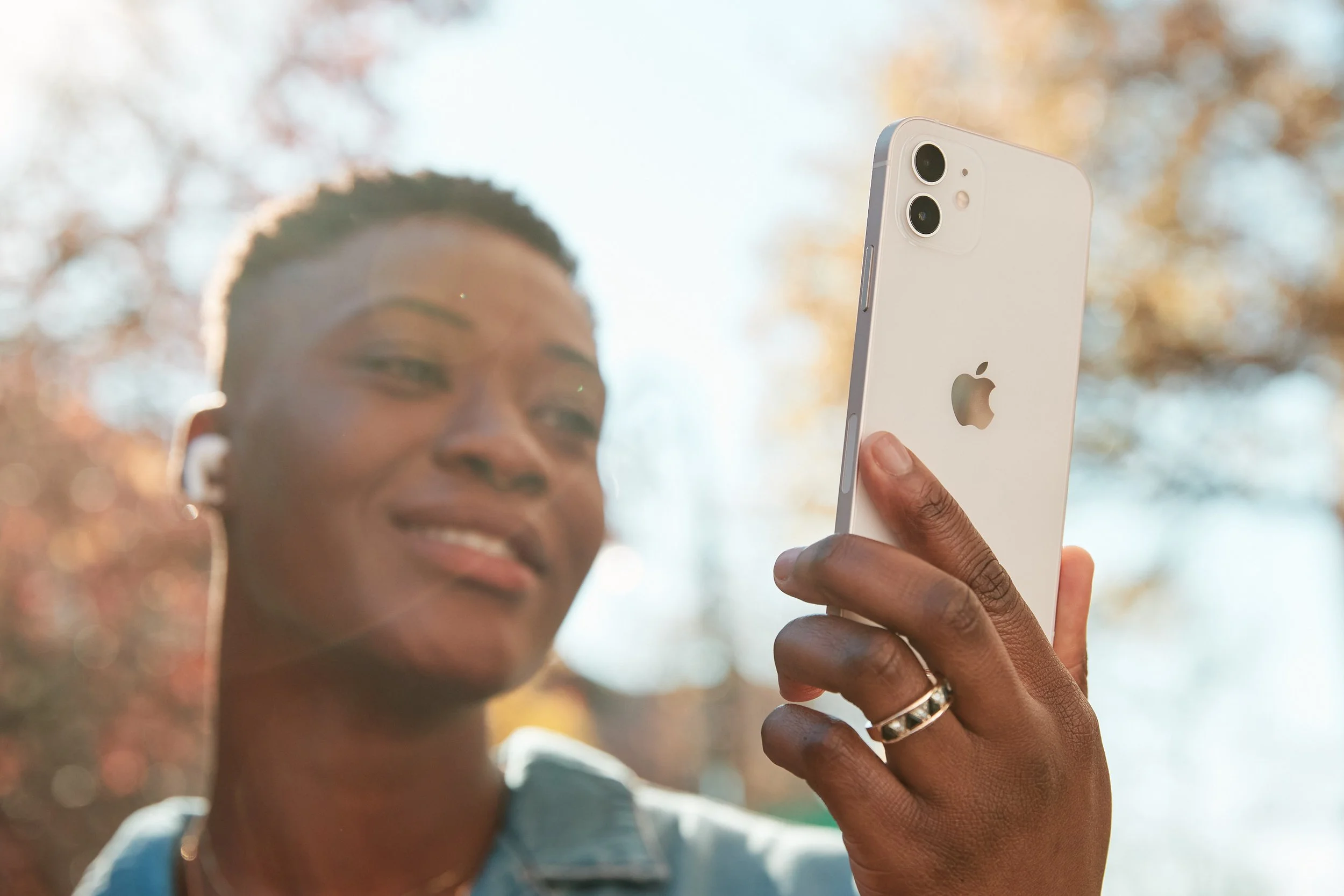 A woman with short hair and earrings taking a selfie with her white iPhone outdoors with trees in the background.