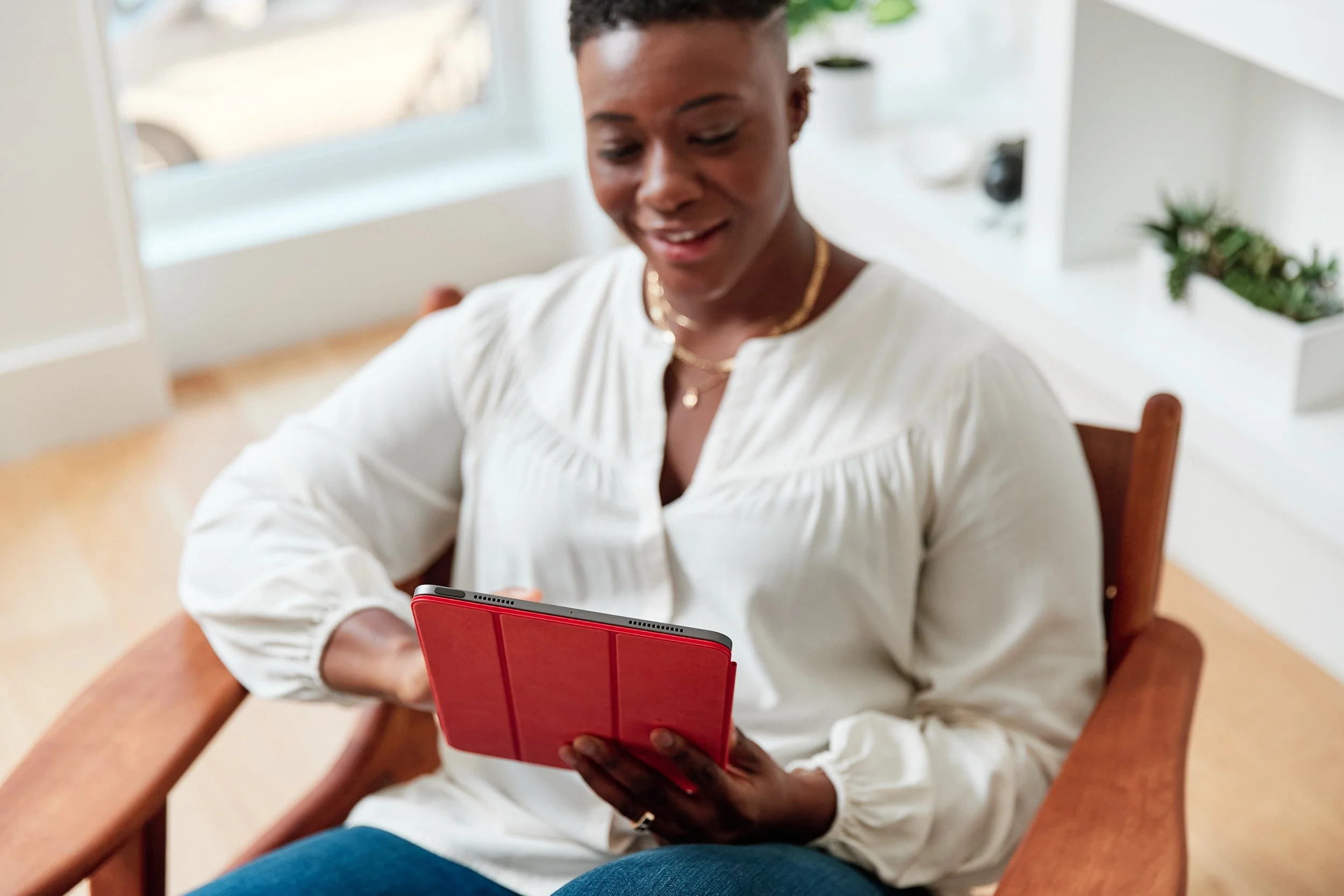 Woman sitting in a wooden chair, looking at her tablet with a red case, indoors near a window with plants on shelves behind her.