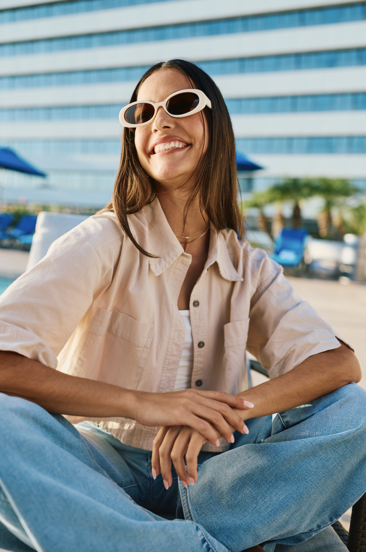 Woman wearing sunglasses smiling while sitting outdoors at a poolside area with a modern high-rise building in the background.