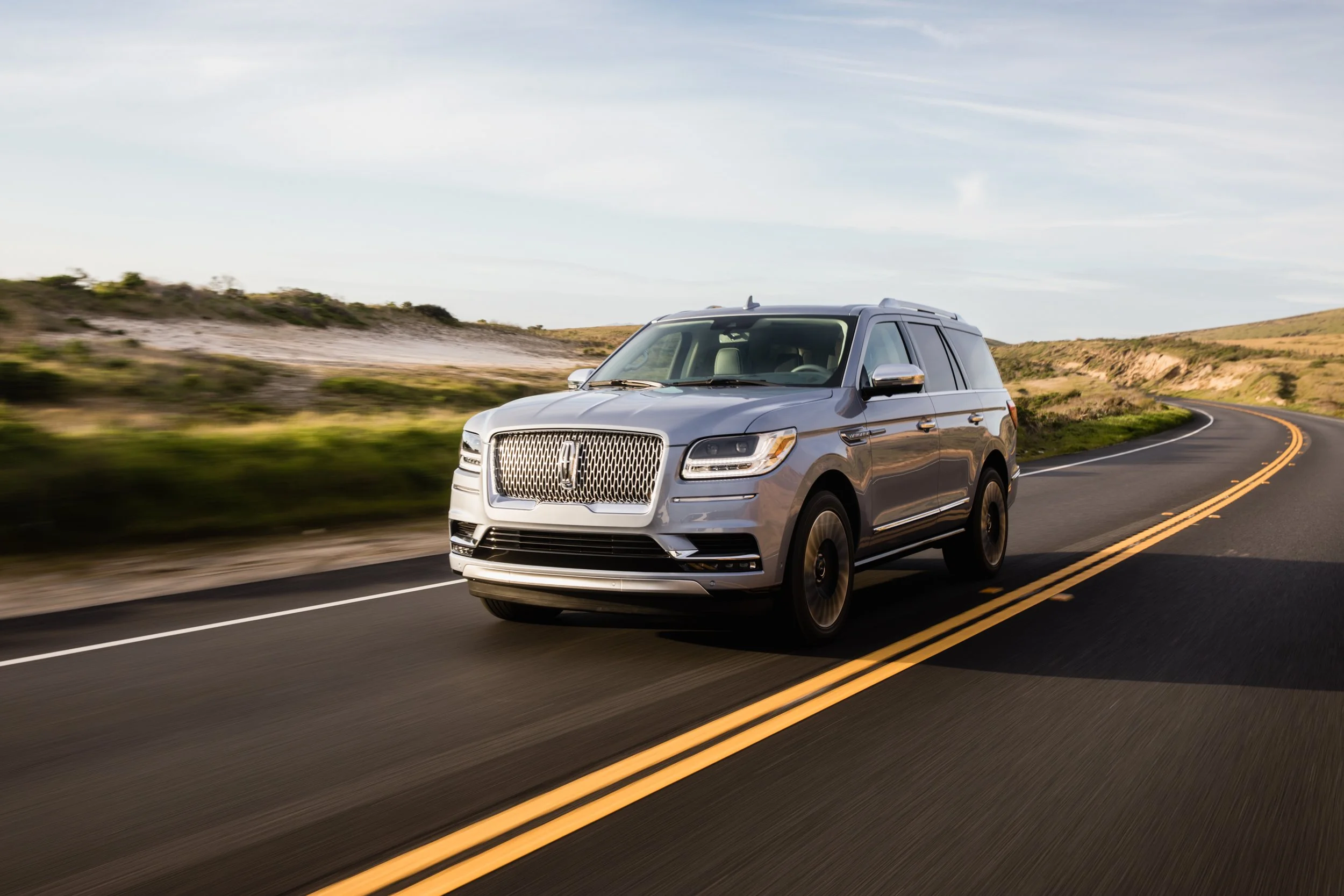 A silver SUV driving on a winding two-lane road in a rural landscape with grassy hills and a partly cloudy sky.