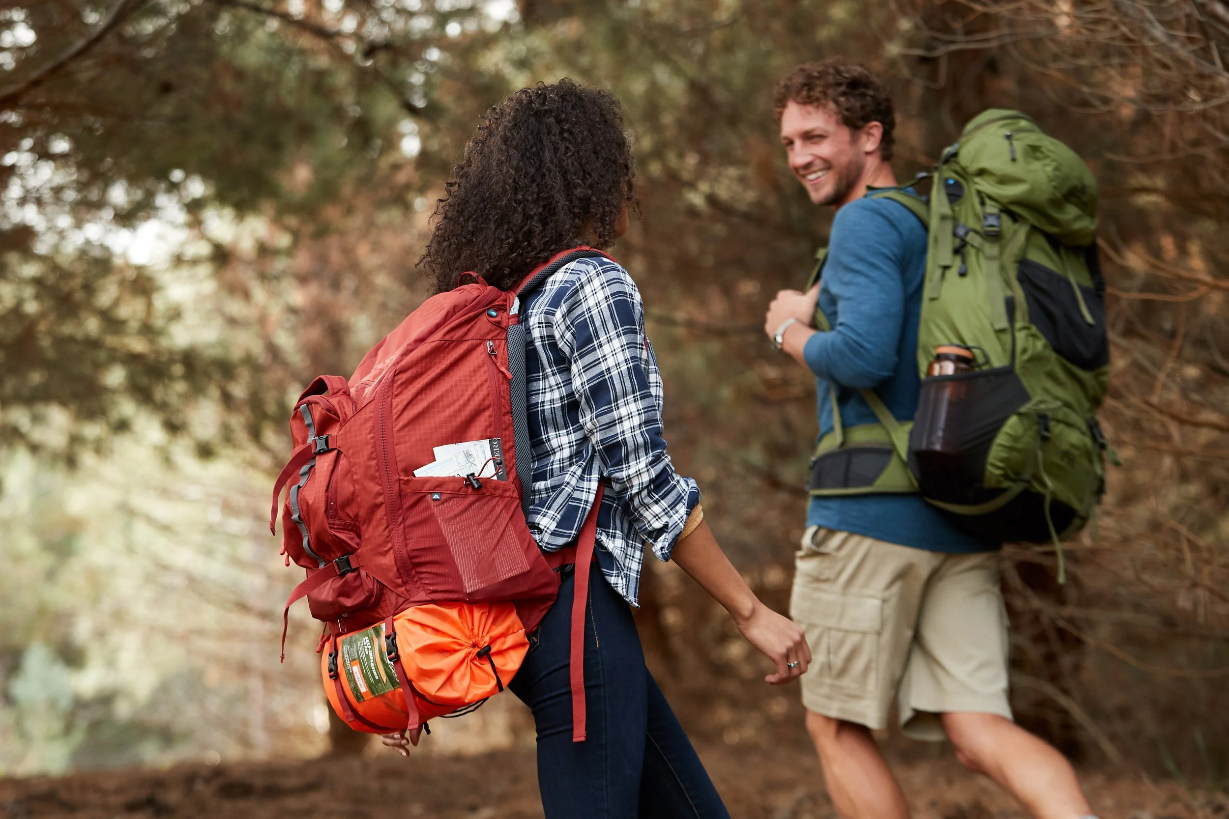 A man and woman hiking with backpacks in a wooded area, smiling and talking to each other.