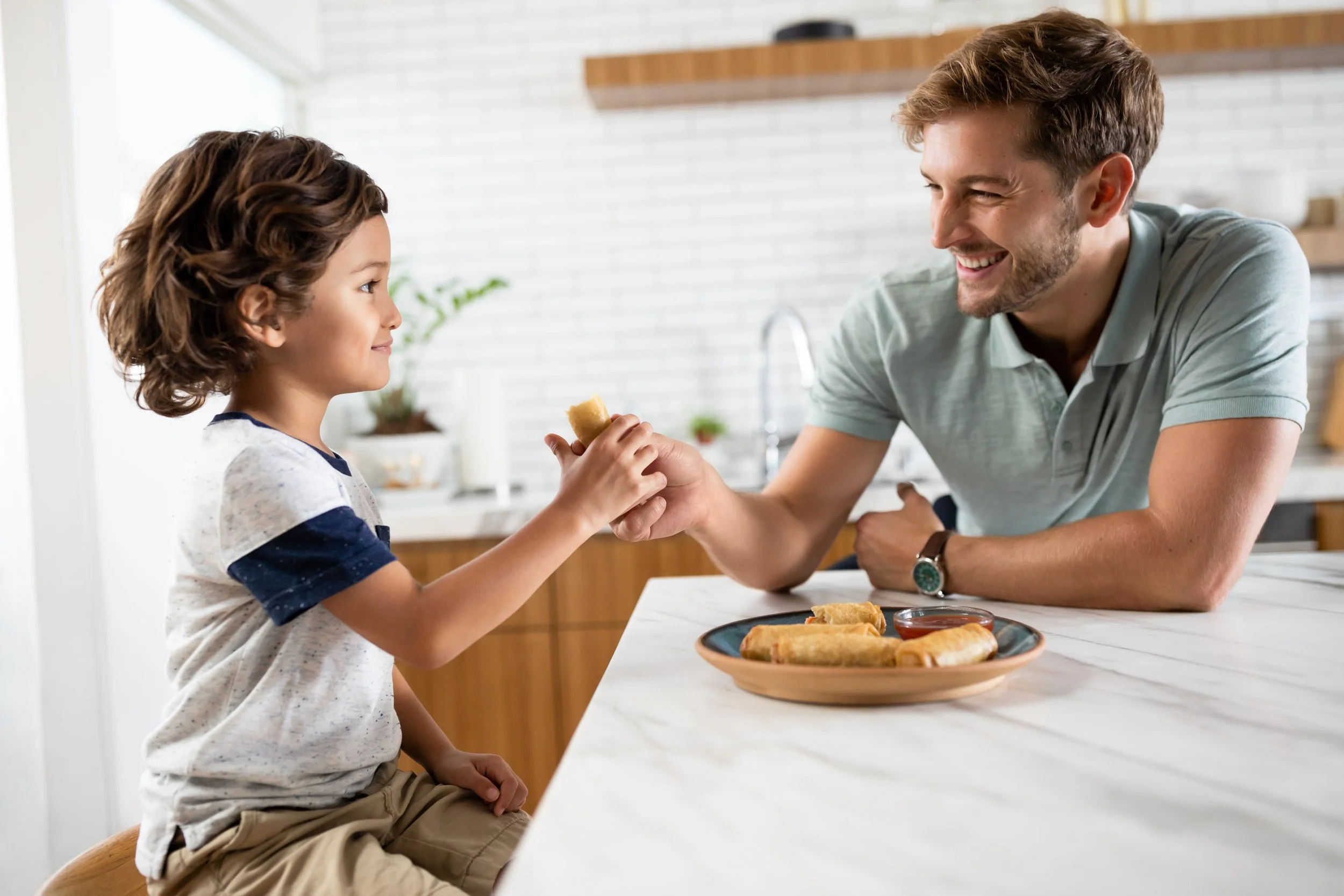 A young boy and a man sharing a handshake at a kitchen counter, with breakfast food on a plate in front of them.
