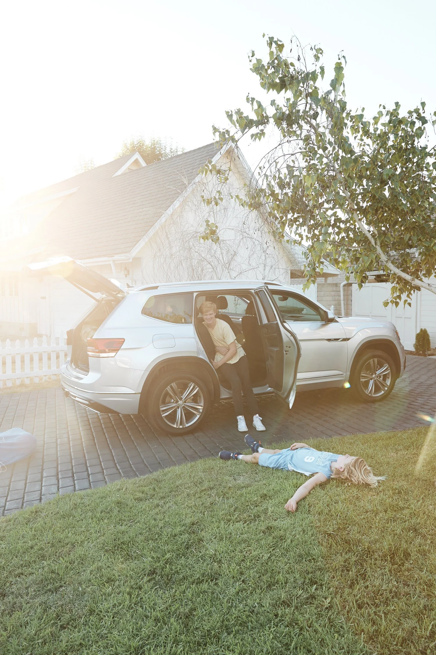 A young boy and girl near a silver SUV parked in a driveway. The girl is smiling and standing by the open car door, while the boy lies on the grass wearing a blue sports jersey. The scene is sunlit with a house, tree, and yard in the background.