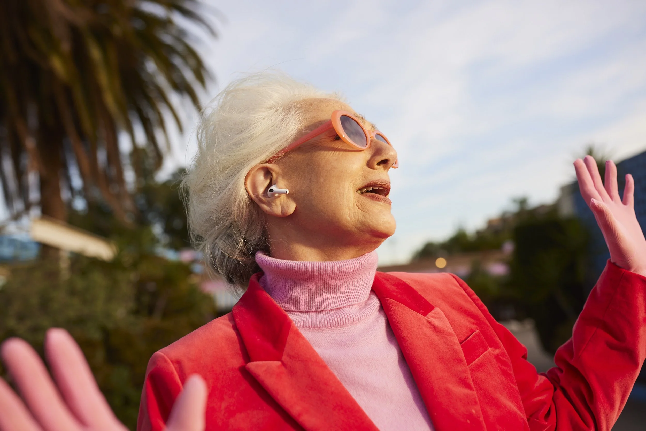 An older woman with white hair wearing sunglasses, pink turtleneck, and red blazer, smiling outdoors with her right hand raised.