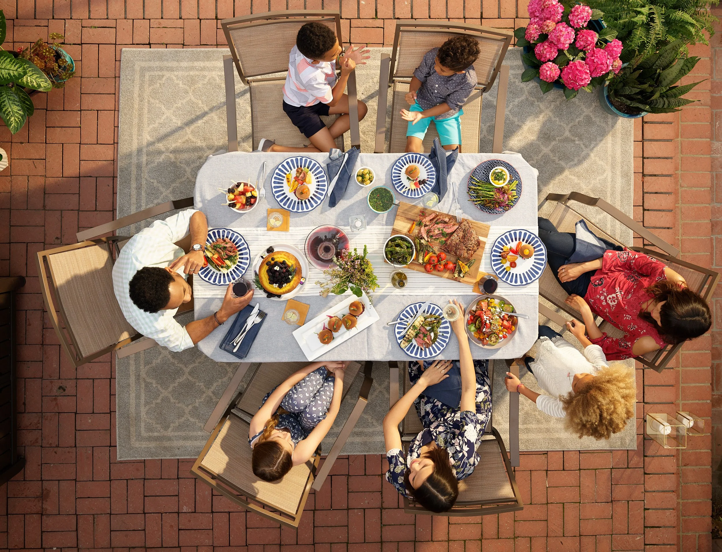 Top-down view of a family dinner with a table filled with various dishes and six people sitting around it in an outdoor patio with brick flooring and potted plants.