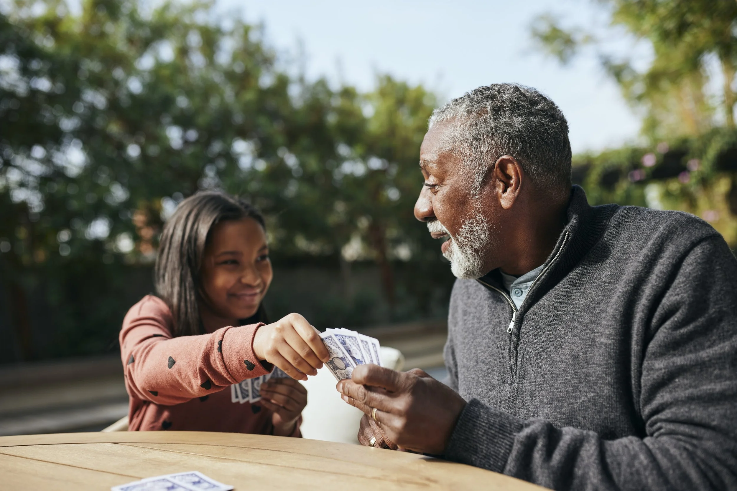 A girl giving dollar bills to an elderly man at a table outdoors with trees in the background.