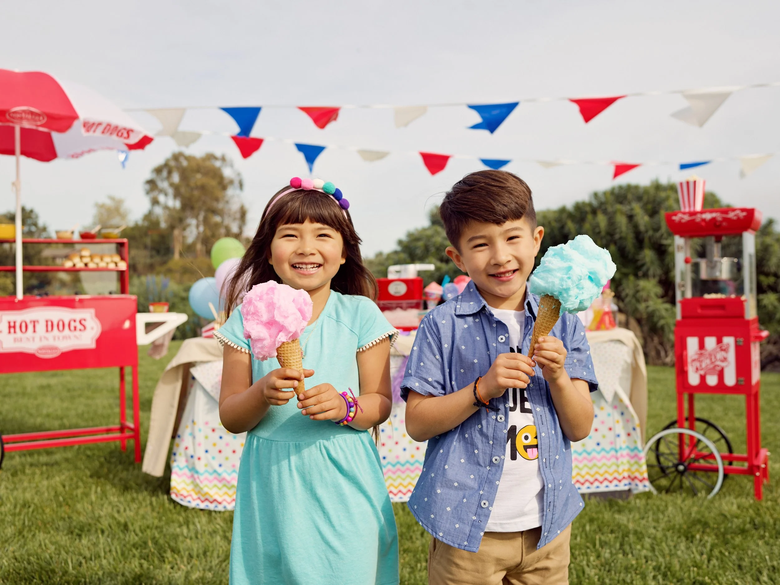 Two children smiling at a fair holding cotton candy over a green grass field with festive decorations and a snack stand in the background.