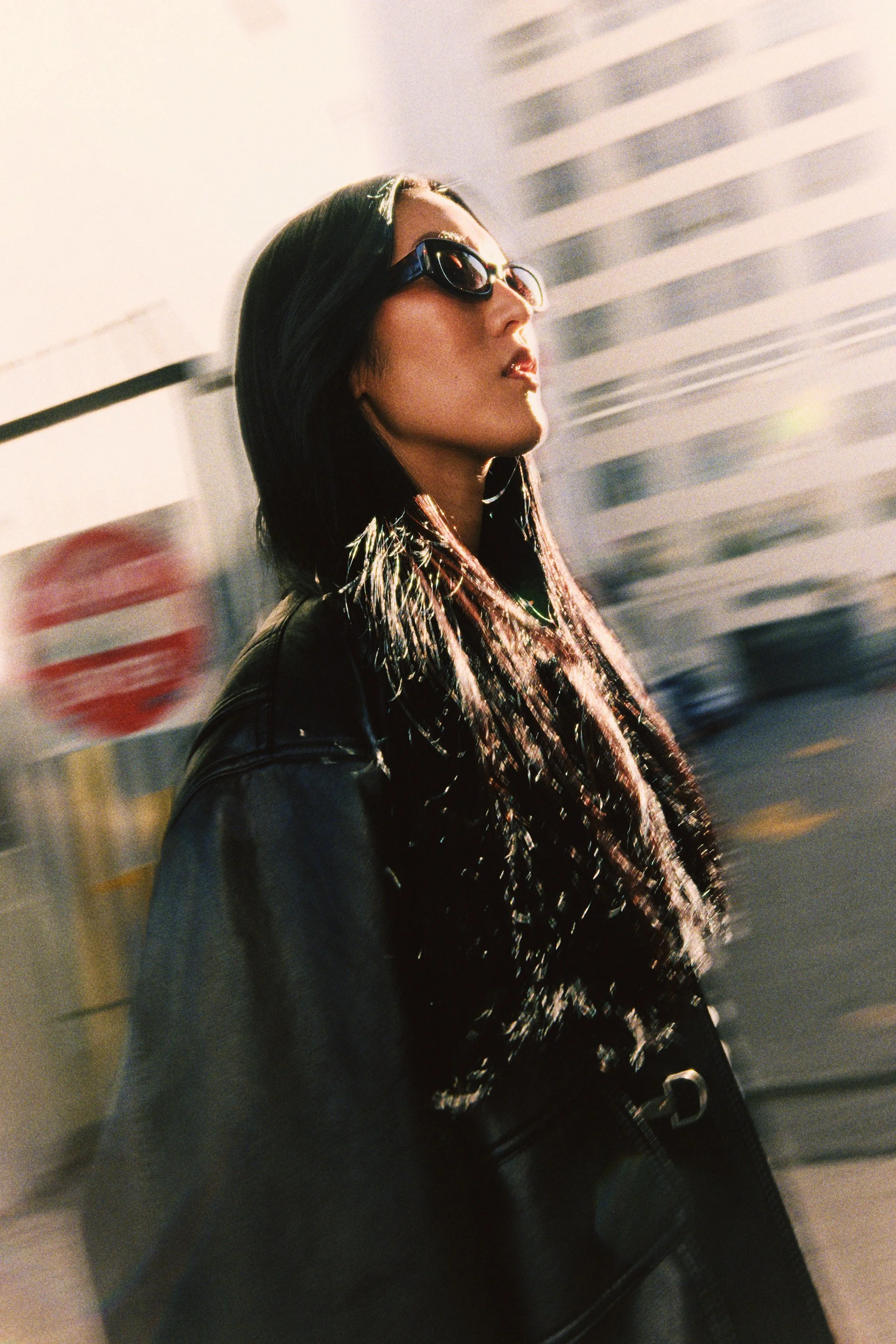 Young woman with dark hair, sunglasses, wearing a black leather jacket, walking outdoors in an urban area with tall buildings in the background.