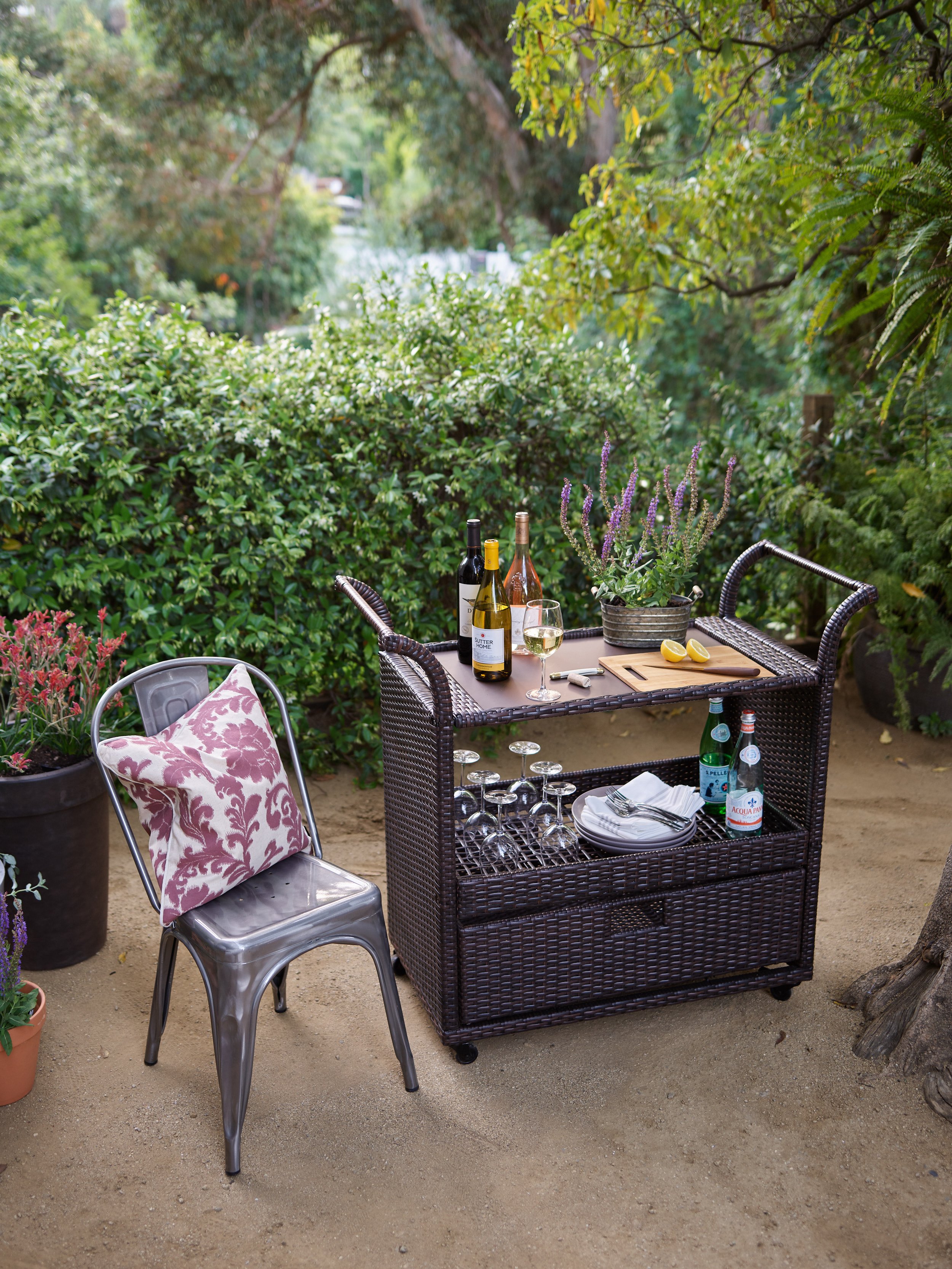 Outdoor patio table with wine bottles, a glass of white wine, a potted plant with purple flowers, a cutting board with lemons, and a wicker storage unit with glasses and bottles, surrounded by bushes and trees.