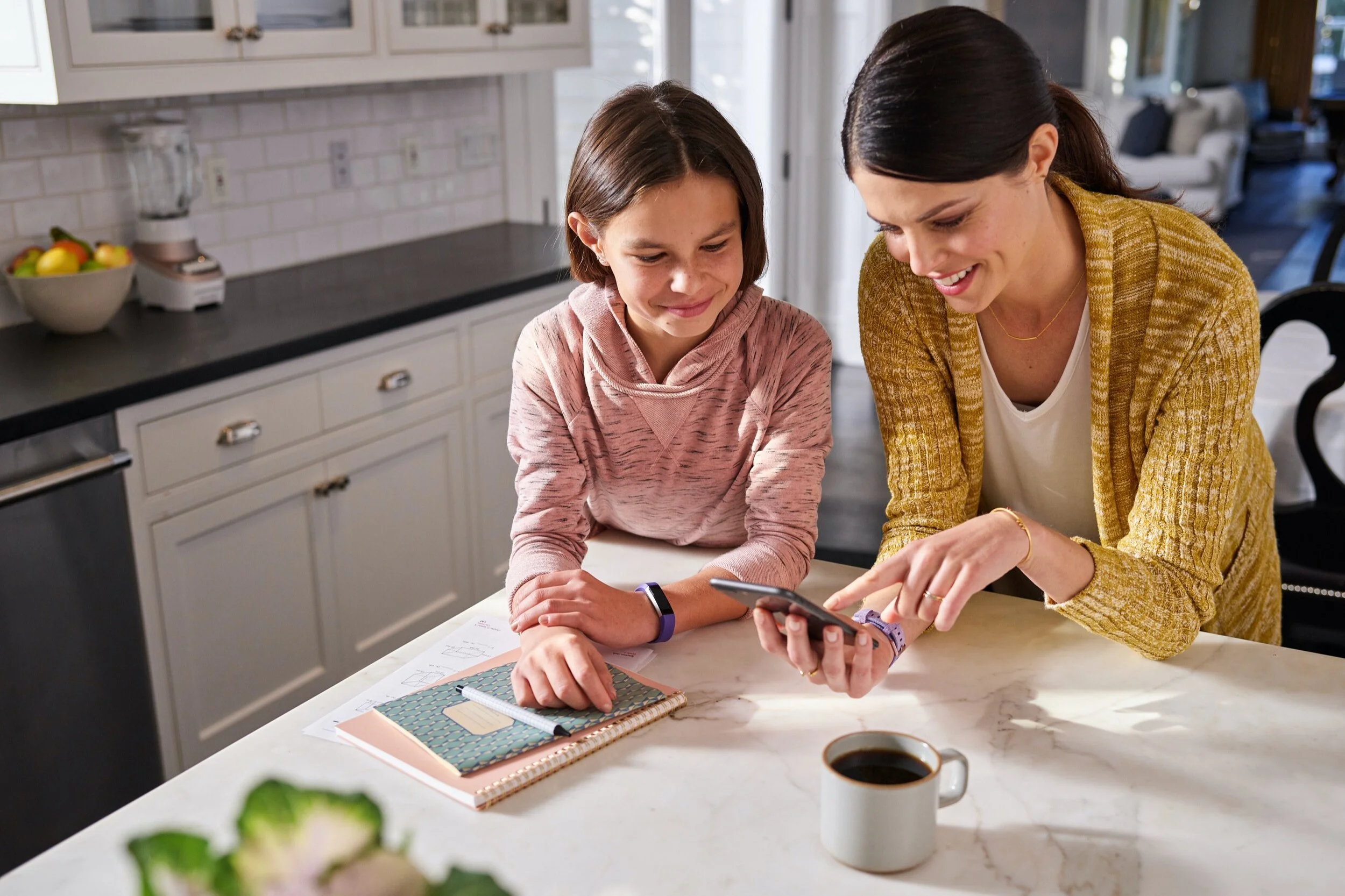 A woman and a girl smiling and looking at a phone together in a kitchen, with a mug of coffee on the counter.