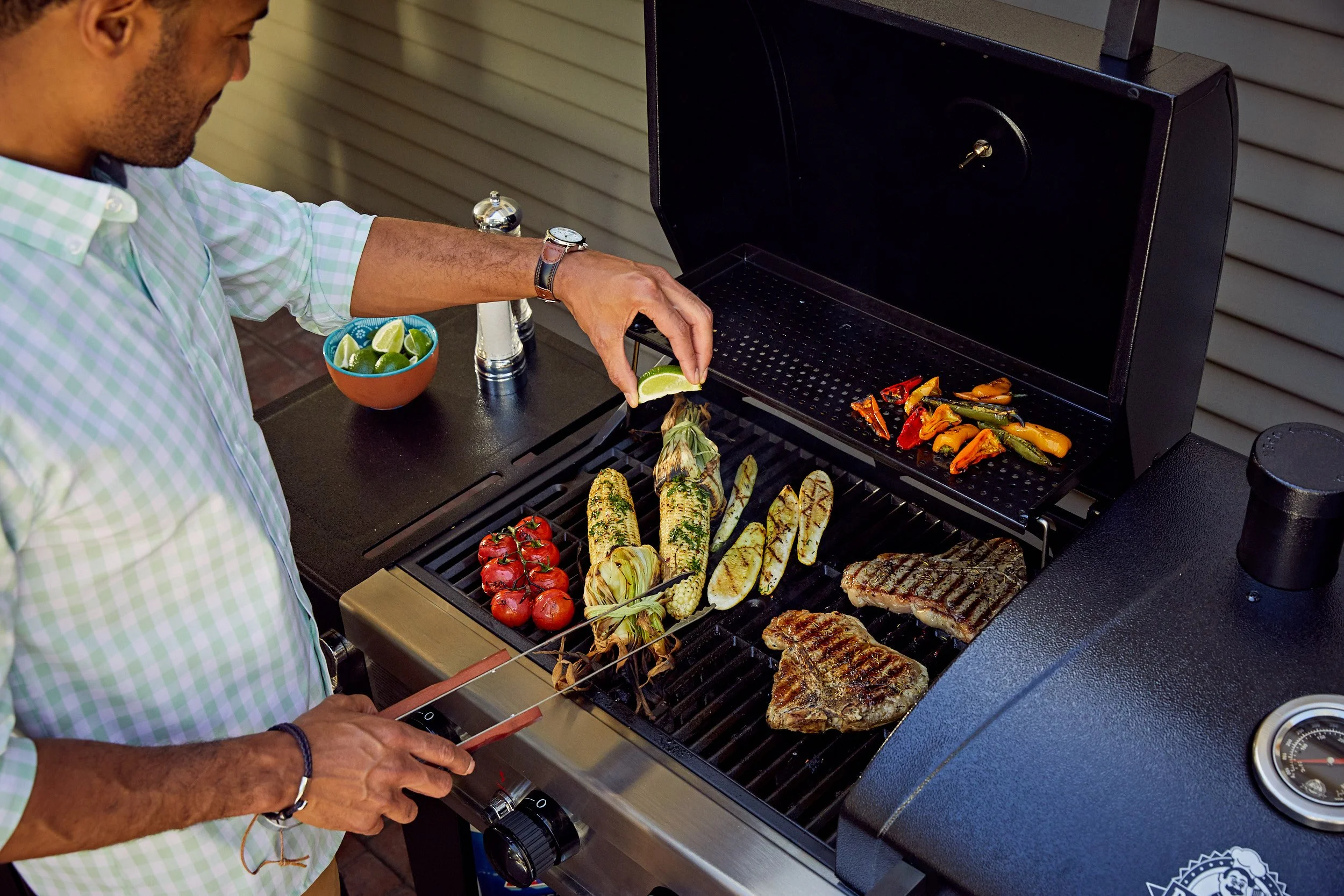 Man grilling vegetables and steaks on a barbecue grill, with cherry tomatoes, zucchini, corn, and mixed peppers, and preparing lime wedges.