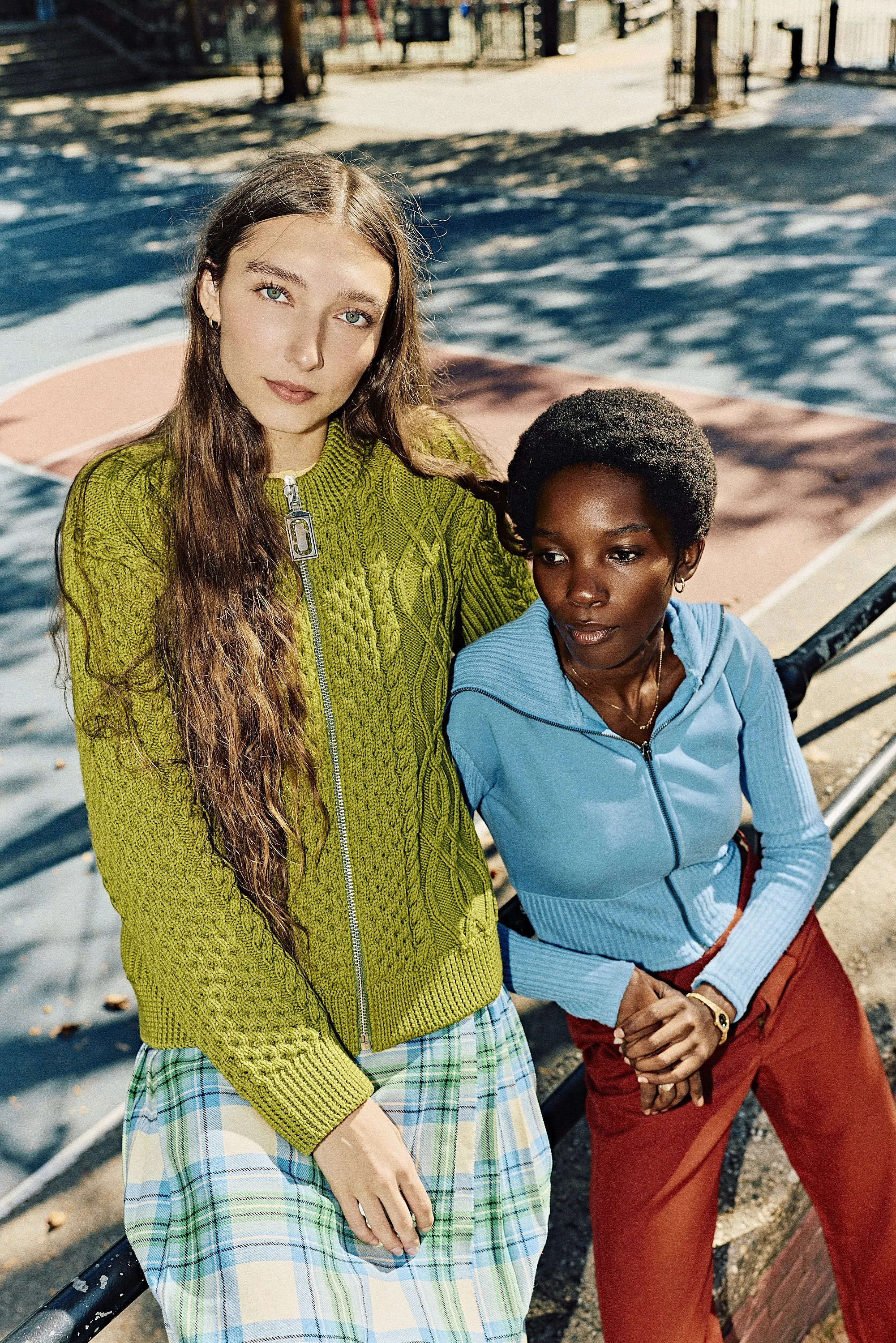 Two women sitting on a park bench next to a basketball court, with trees and a fence in the background, enjoying a sunny day.