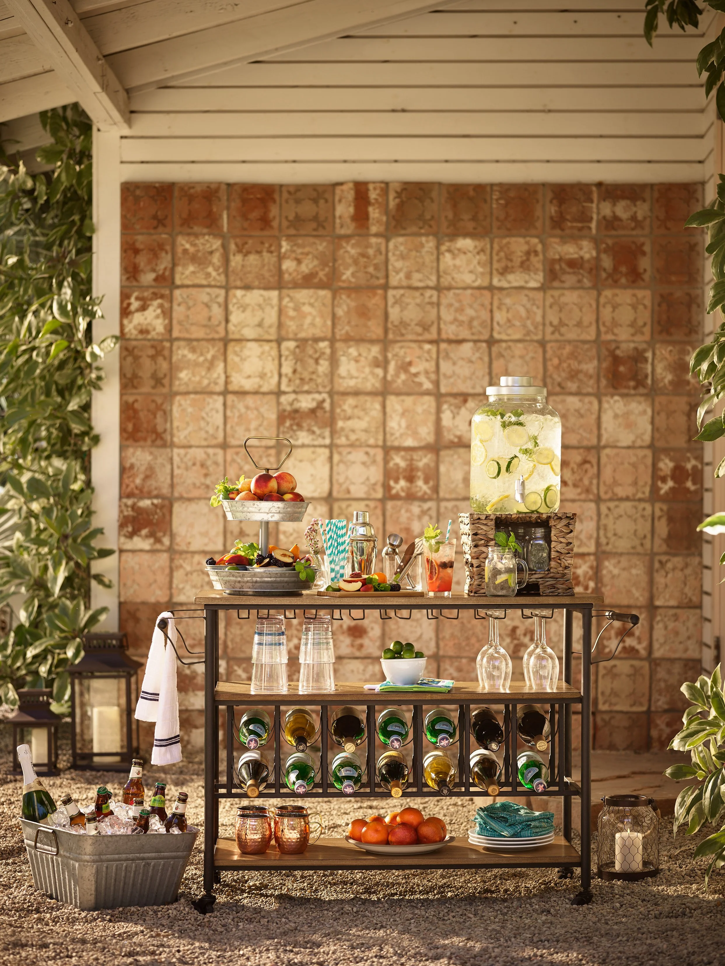 Outdoor drink and food station with a black metal cart holding glass bottles of beer, wine, plates of apples, and decorative cups, decorated with greenery and lanterns in a backyard setting.