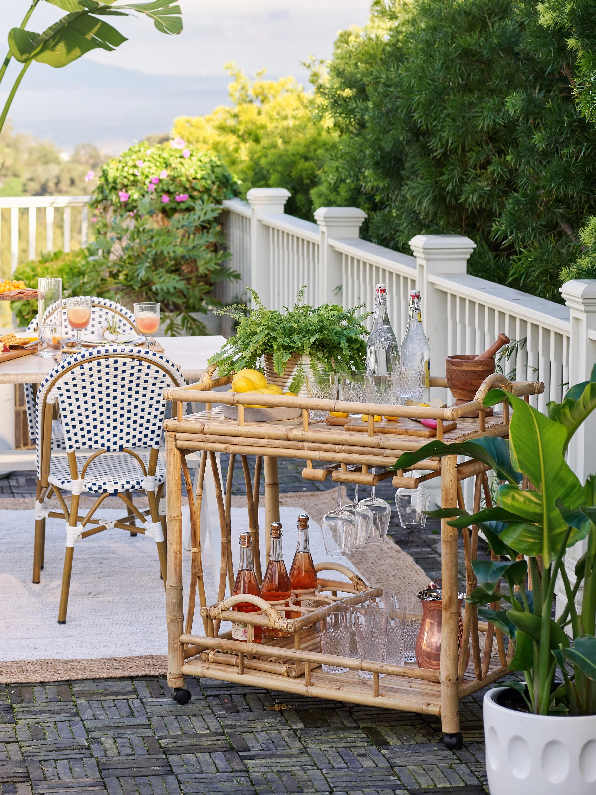 Outdoor patio with a bamboo bar cart holding drinks, glasses, and lemons, nearby a table with drinks and snacks, surrounded by green plants and a view of trees and sky.