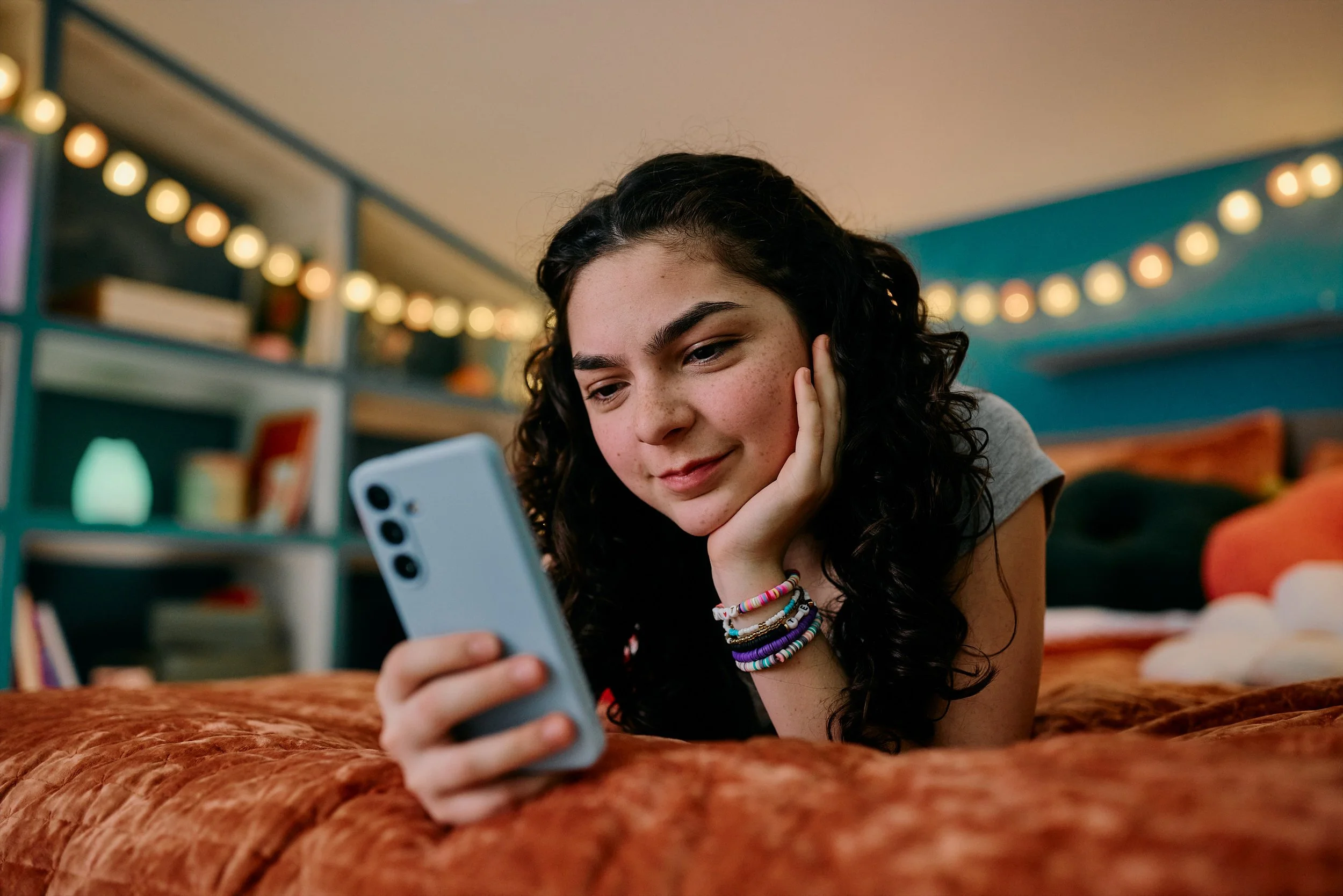A young girl with dark curly hair, multiple colorful bracelets, and freckles, lying on her stomach on a brown plush bed, looking at her smartphone with a slight smile, in a cozy room decorated with string lights and shelves filled with books and deco