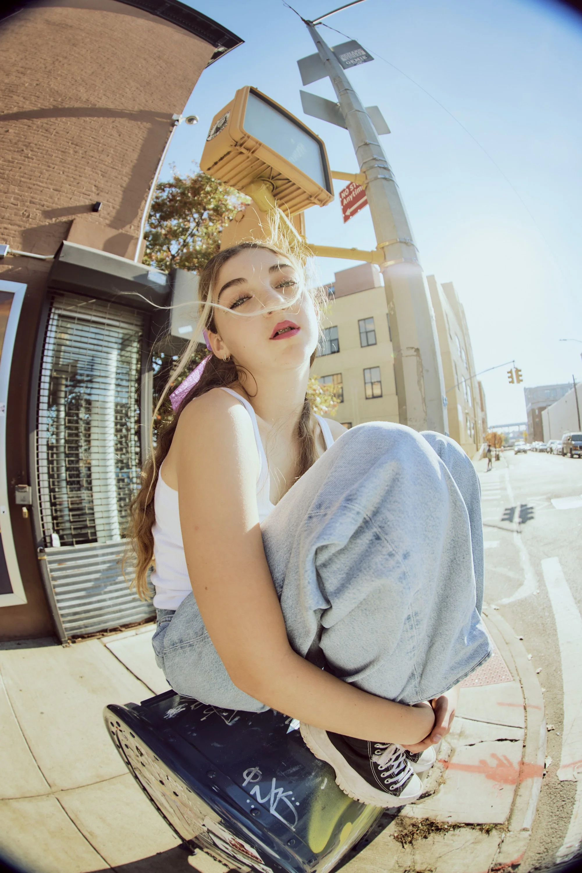 Young woman with long hair sitting on a black crate on a city sidewalk, with an overexposed sky, buildings, and street signs in the background.
