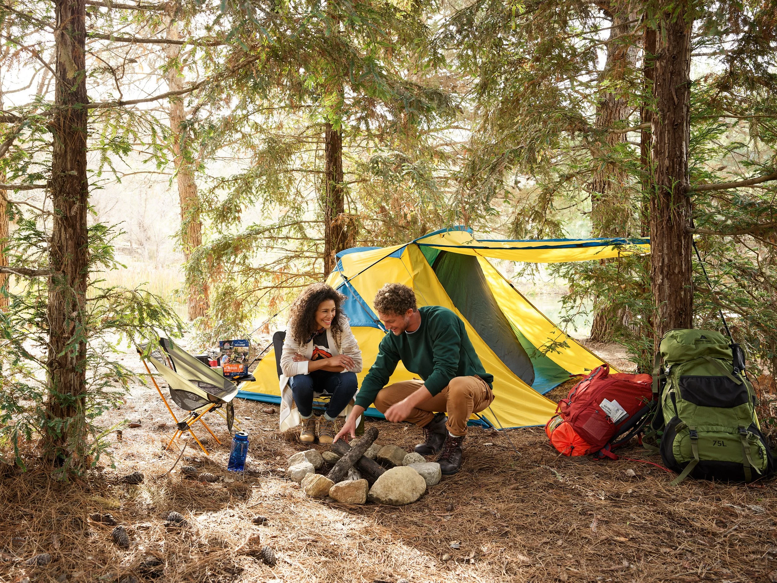 A man and woman sitting around a campfire in a wooded area with a yellow and blue tent, backpacks, and camping gear nearby.