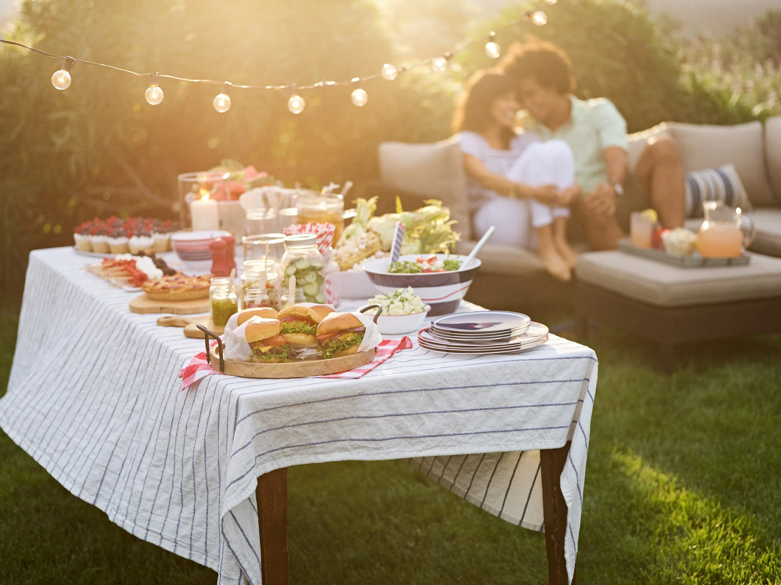 Outdoor summer gathering with a feast on a striped tablecloth, including sandwiches, salads, desserts, and drinks, with a couple sitting on a couch in the background.