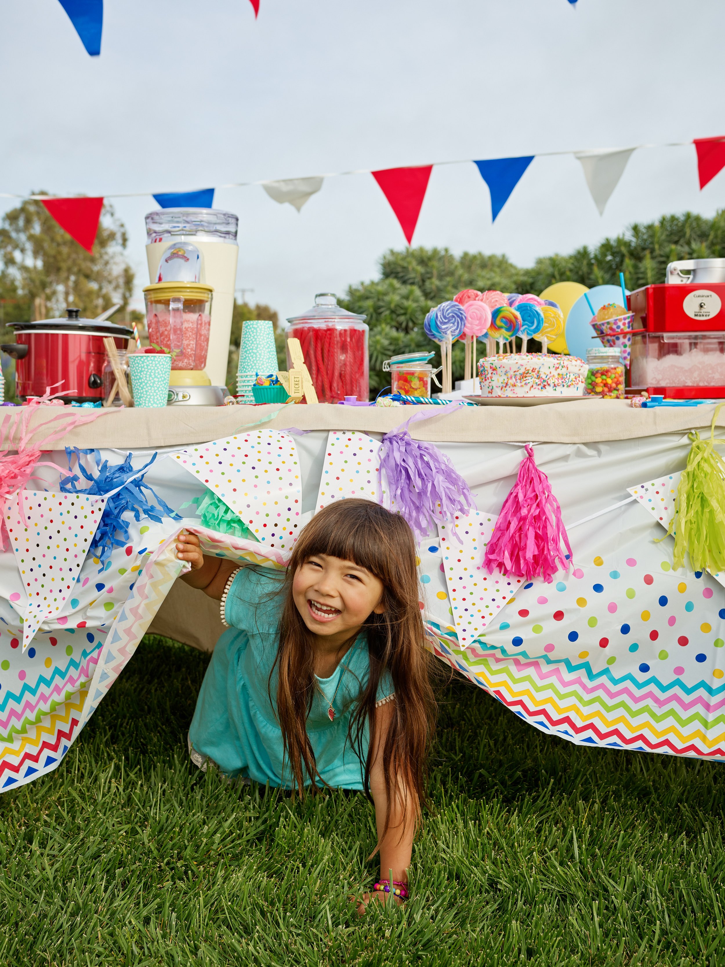 Happy girl peeking out from underneath a decorated outdoor table at a colorful birthday party, with food, cake, and decorations on the table.