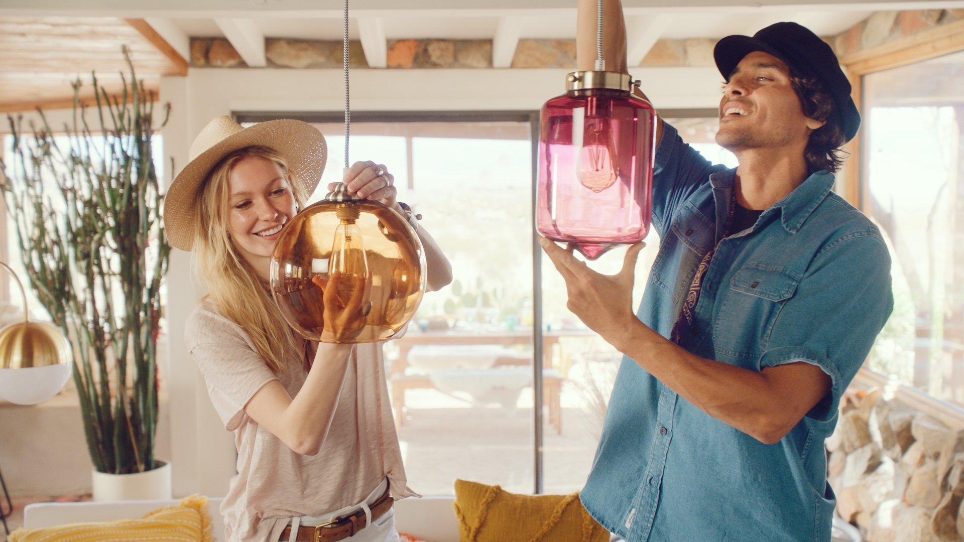 Two people, a woman and a man, installing pendant lights in a bright living room with large windows, a stone accent wall, and cozy furniture.