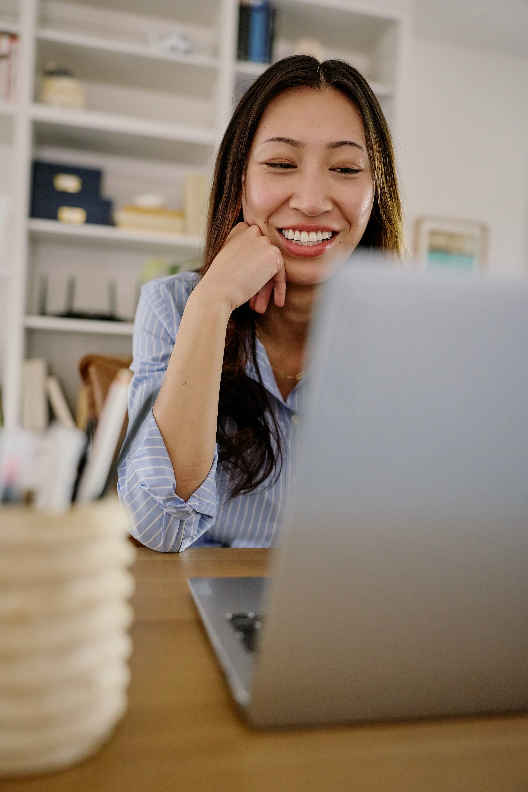 A woman with long dark hair smiling while looking at her laptop screen in a home office setting.