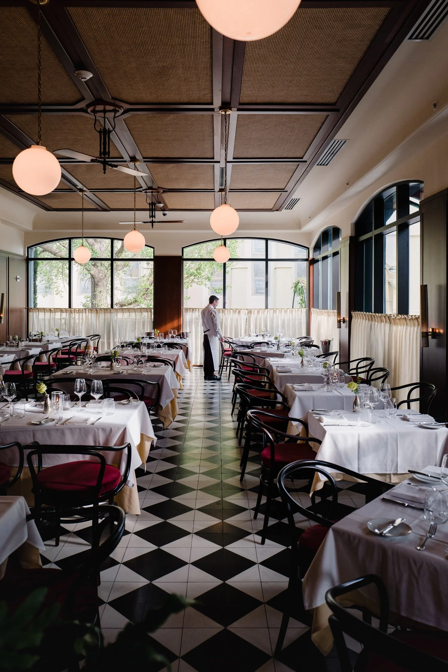 Elegant restaurant dining area with white tablecloths, wine glasses, and chairs, with a waiter standing by the window.