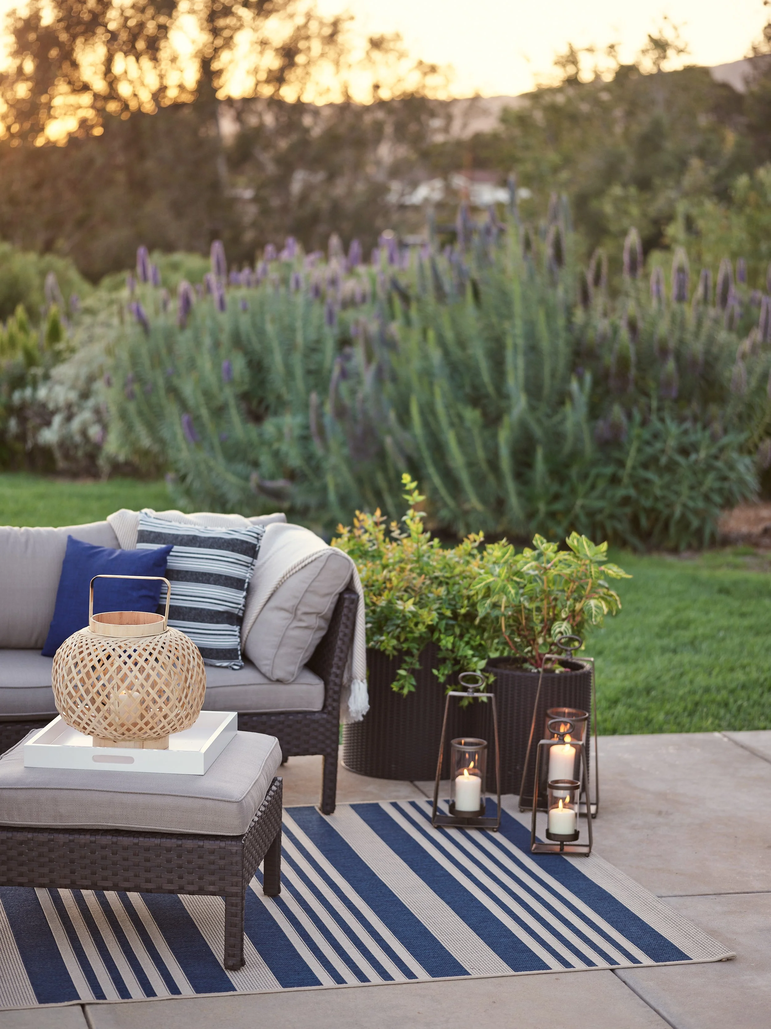 Outdoor patio setup during sunset with a beige cushioned outdoor sofa, striped blue and white pillows, a matching ottoman with a decorative lantern, a potted plant, and three candle lanterns on the ground. In the background, there are tall purple flo