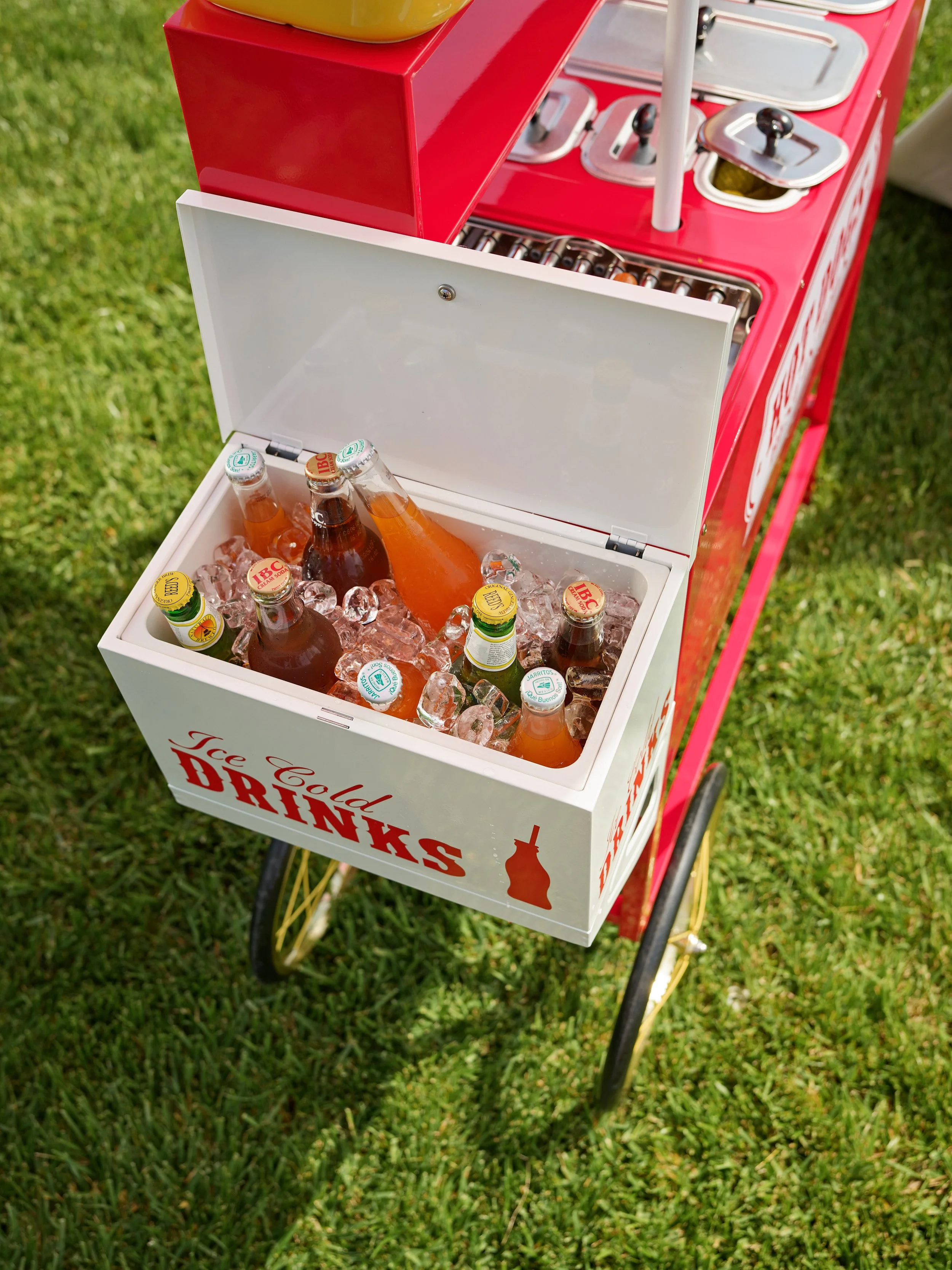 Ice cooler filled with bottles of soda and iced drinks on a red beverage cart outdoors.