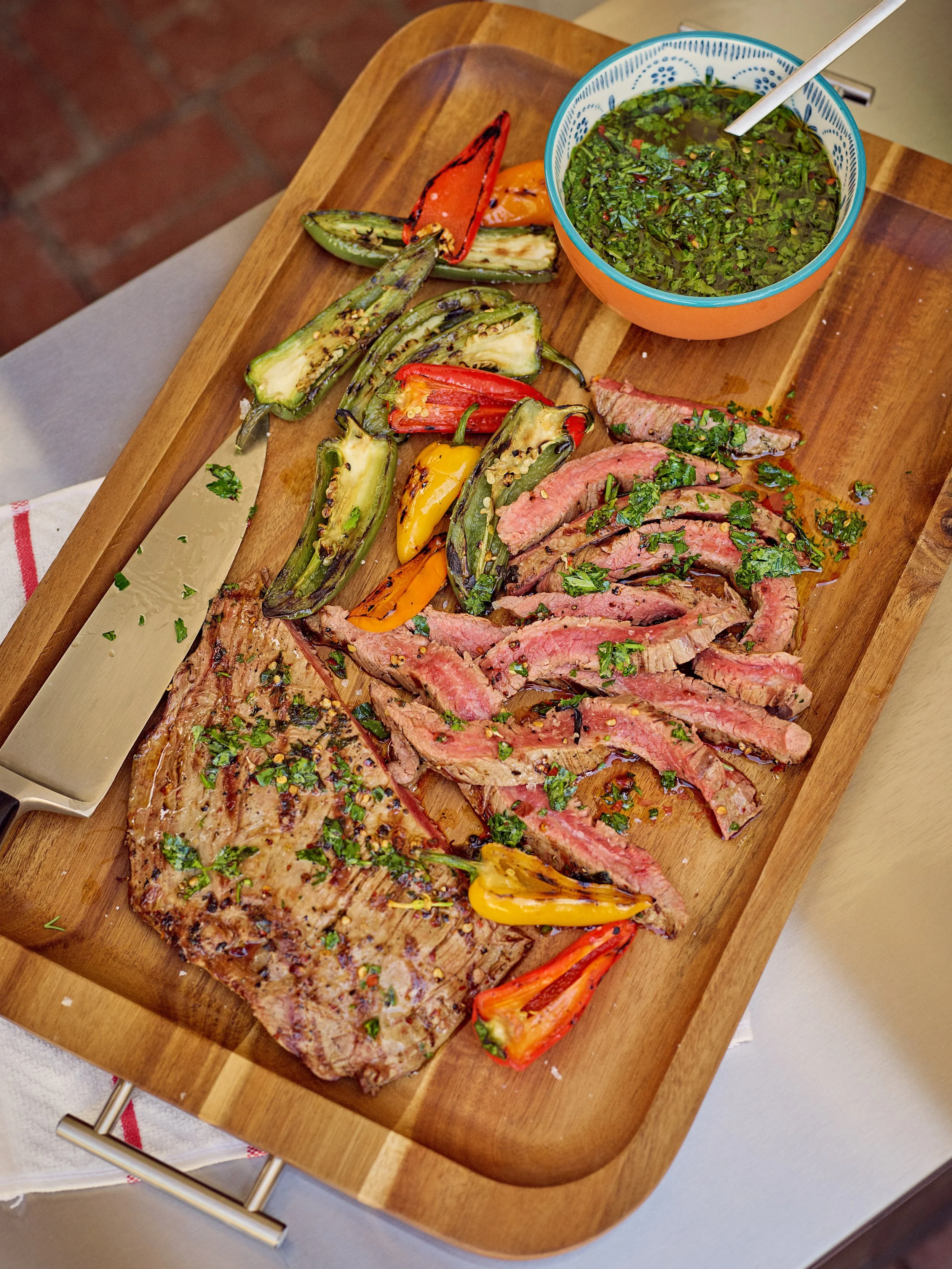 Grilled steak and sliced beef with roasted colorful peppers, a bowl of green sauce, and a steak knife on a wooden serving board.