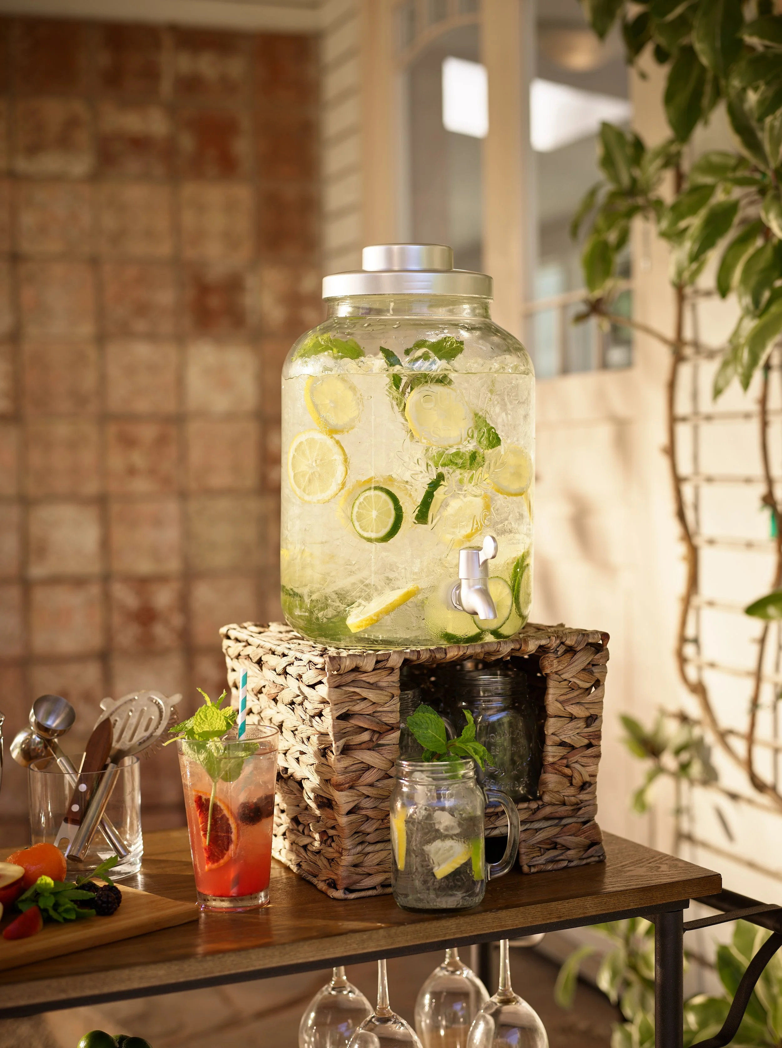 A large glass jar of infused water with lemon, lime, and mint slices sits on a woven stand, with a glass of pink drink beside it and fresh fruit on a wooden table.