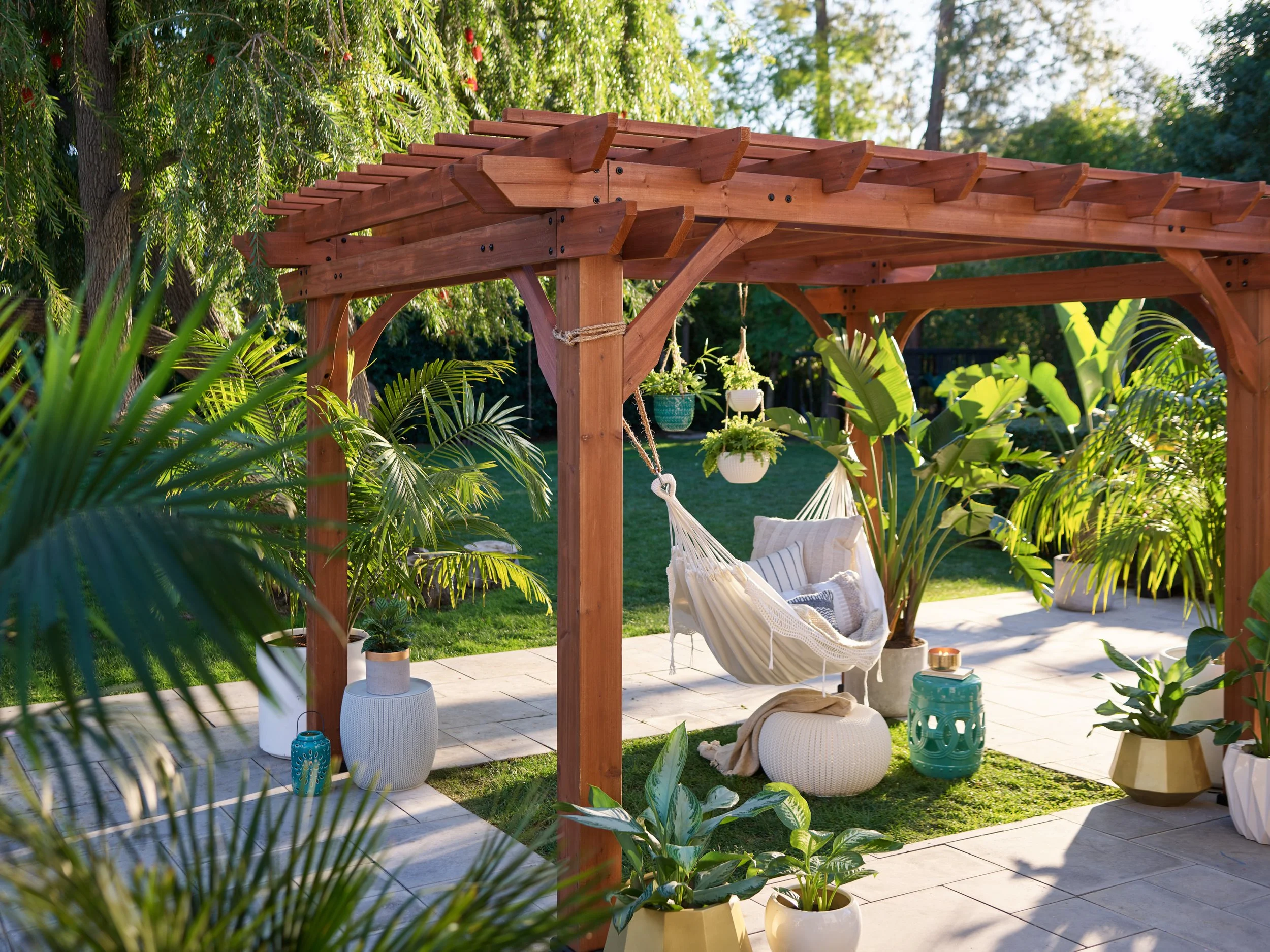 A backyard patio with a wooden pergola, hanging plants, a hammock with cushions, surrounding potted plants, and lush greenery.