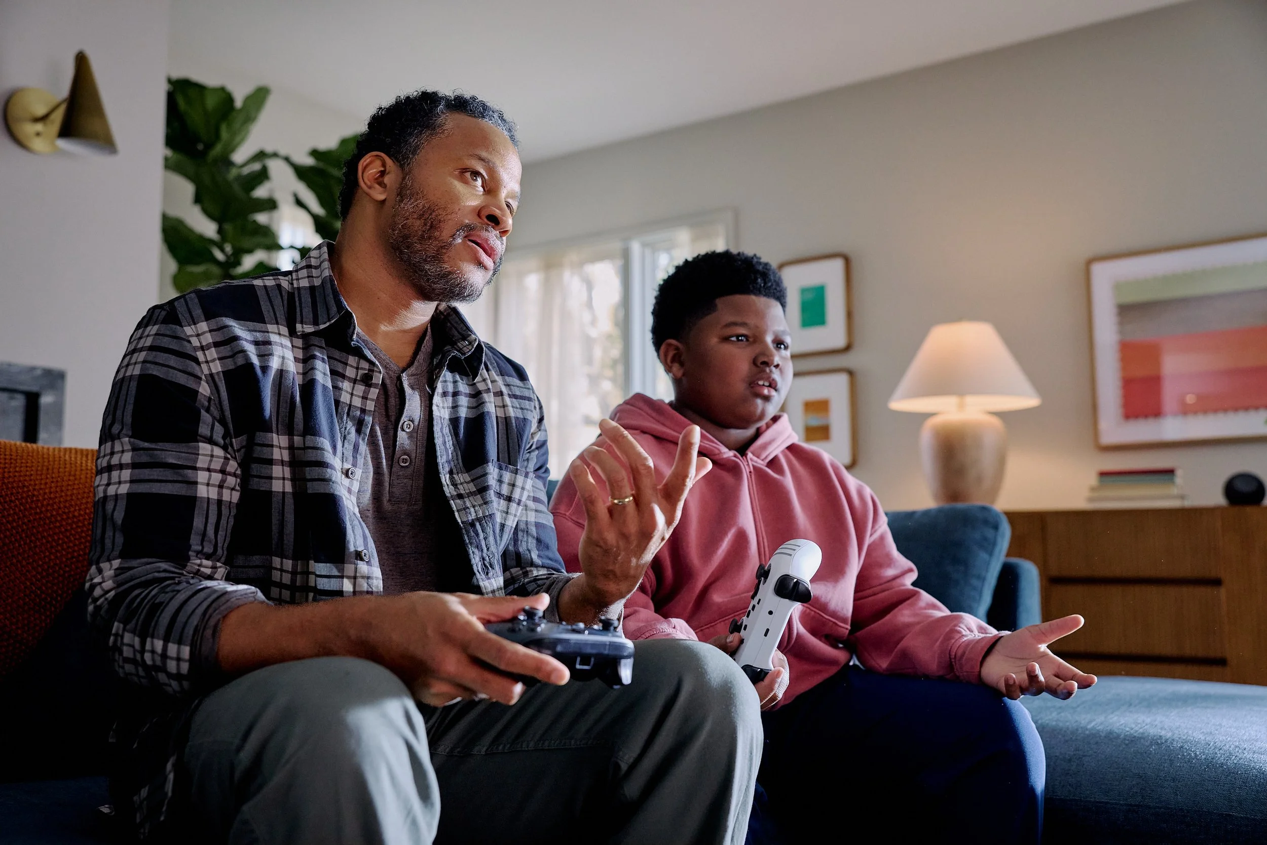An adult man and a young boy sitting on a sofa are playing video games in a living room. The man is wearing a plaid shirt, and the boy is in a pink hoodie, holding two game controllers. They appear to be engaged with the game, with the man's hand ges