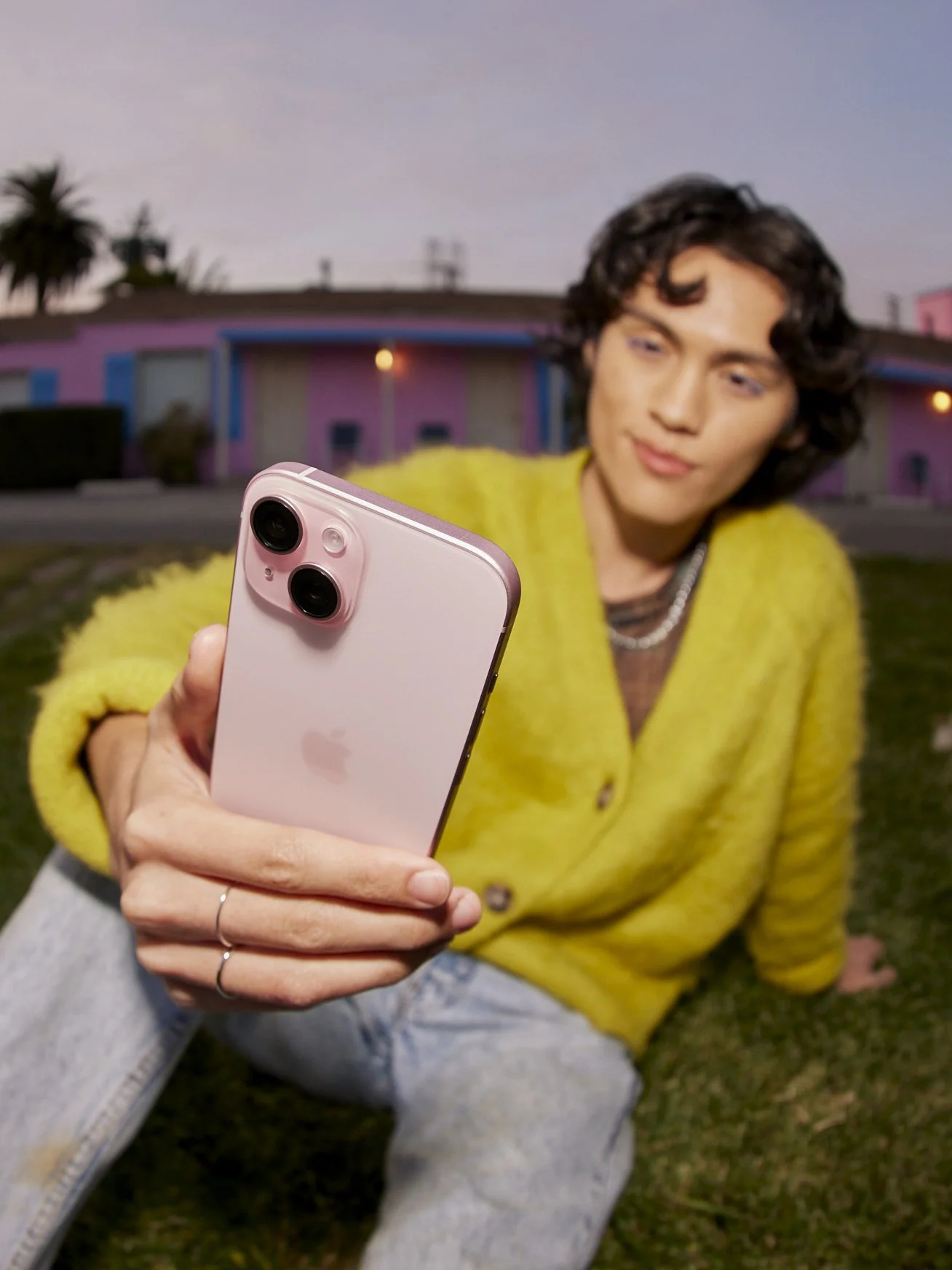 Person in a yellow cardigan taking a selfie with a pink iPhone in front of a pink house with blue accents during sunset.