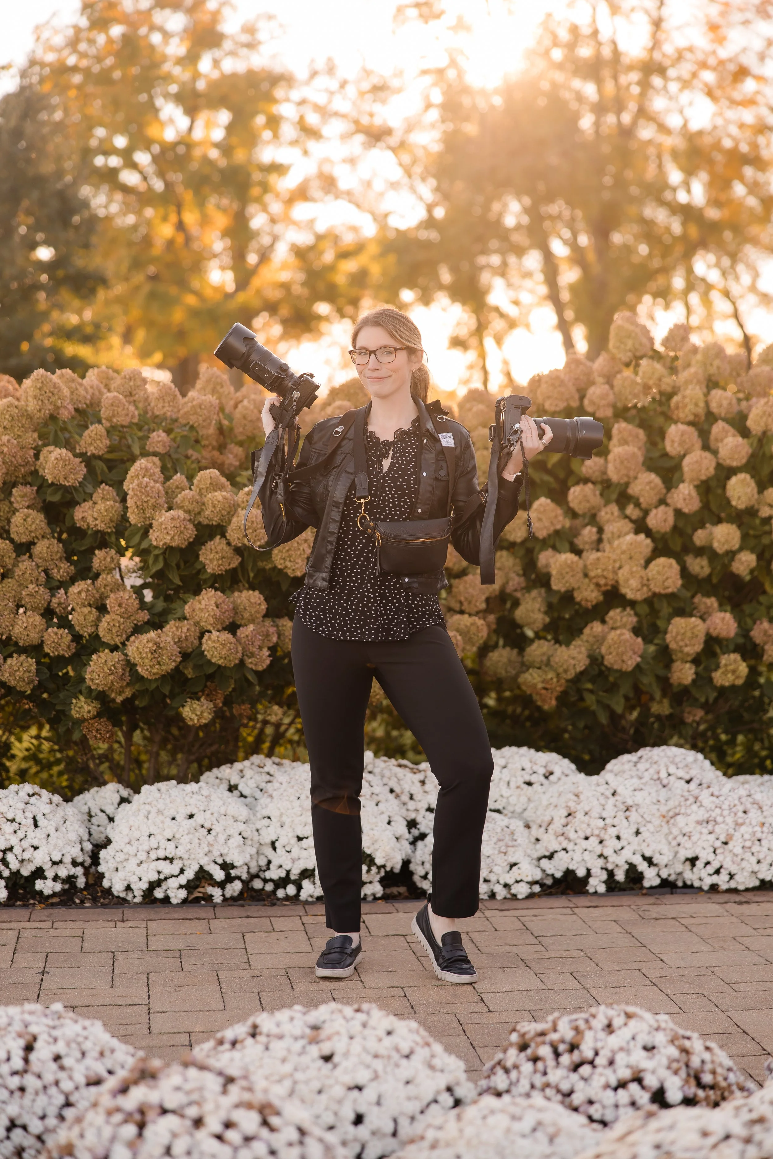 a photo of carol deanda on a wedding day in front of flowers holding two nikon cameras