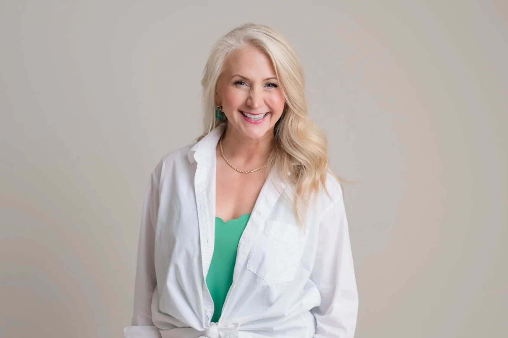 A smiling blonde woman with wavy hair wearing a white shirt tied at the waist, green top, gold necklace, and green earrings against a plain gray background.