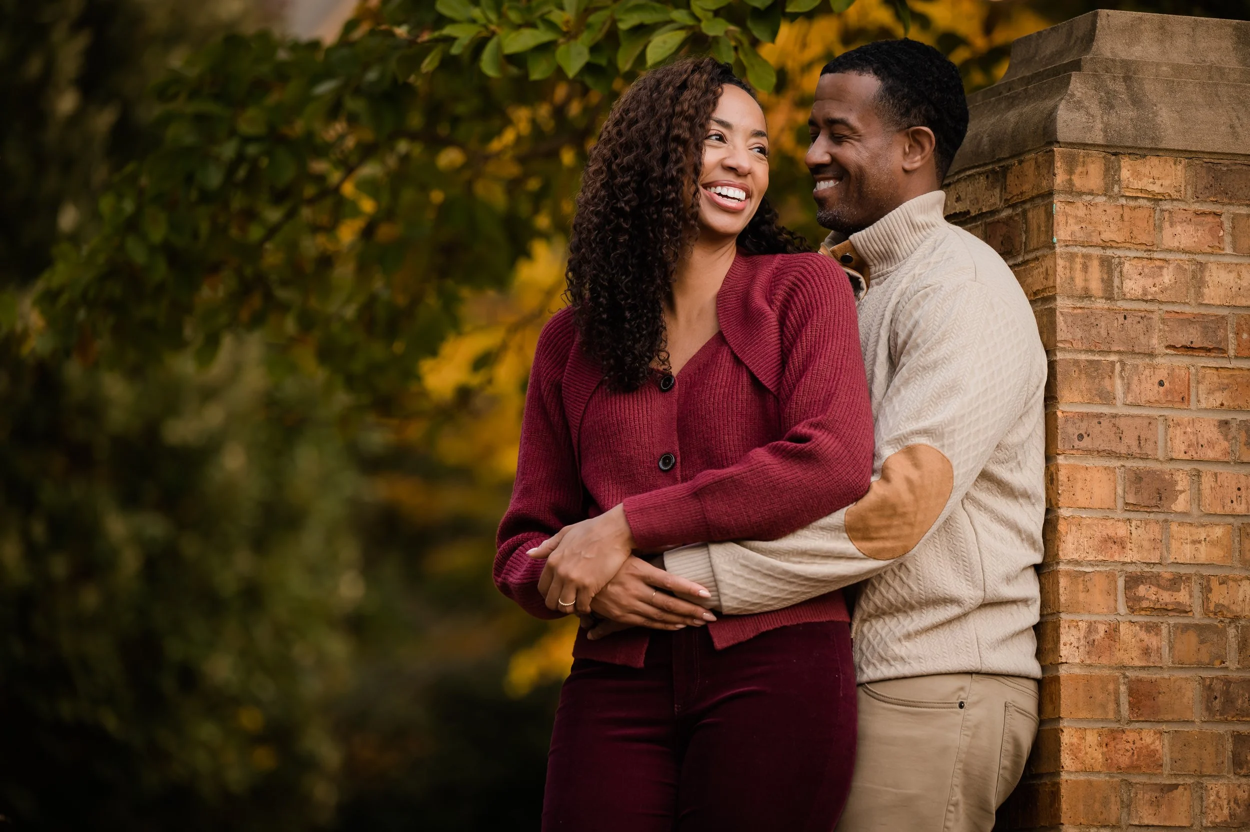 A happy couple, a woman in a red cardigan and a man in a beige sweater, sharing a joyful moment outdoors next to a brick wall with fall foliage in the background.