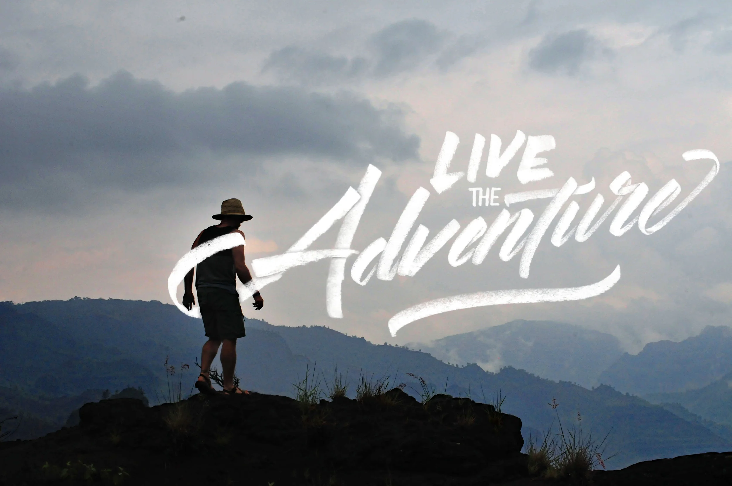Silhouette of a person with a hat standing on a mountain, with a cloudy sky and mountain ranges in the background, and the words 'Live the Adventure' written across the image.